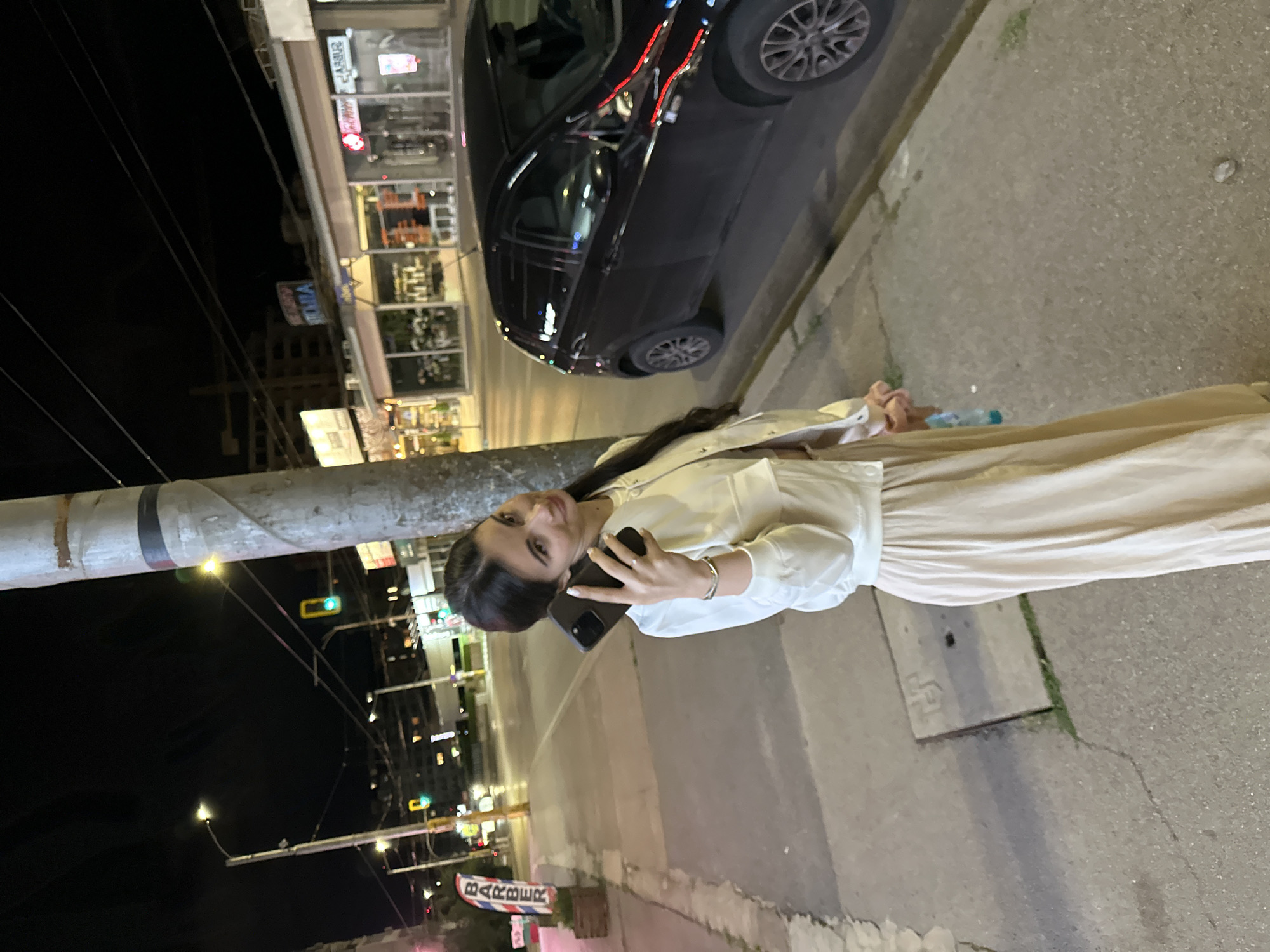 A woman with dark hair is standing on a sidewalk at night, holding a dark-colored smartphone to her ear. She is wearing a light-colored jacket over a beige dress or skirt. To her right, a dark-colored hatchback car is parked along the curb. In the background, a street is visible with traffic lights, streetlights, and buildings with illuminated storefronts. A sign with the word "BARBER" in vertical lettering is positioned to the left of the woman. The scene appears to be a commercial or urban area at night.