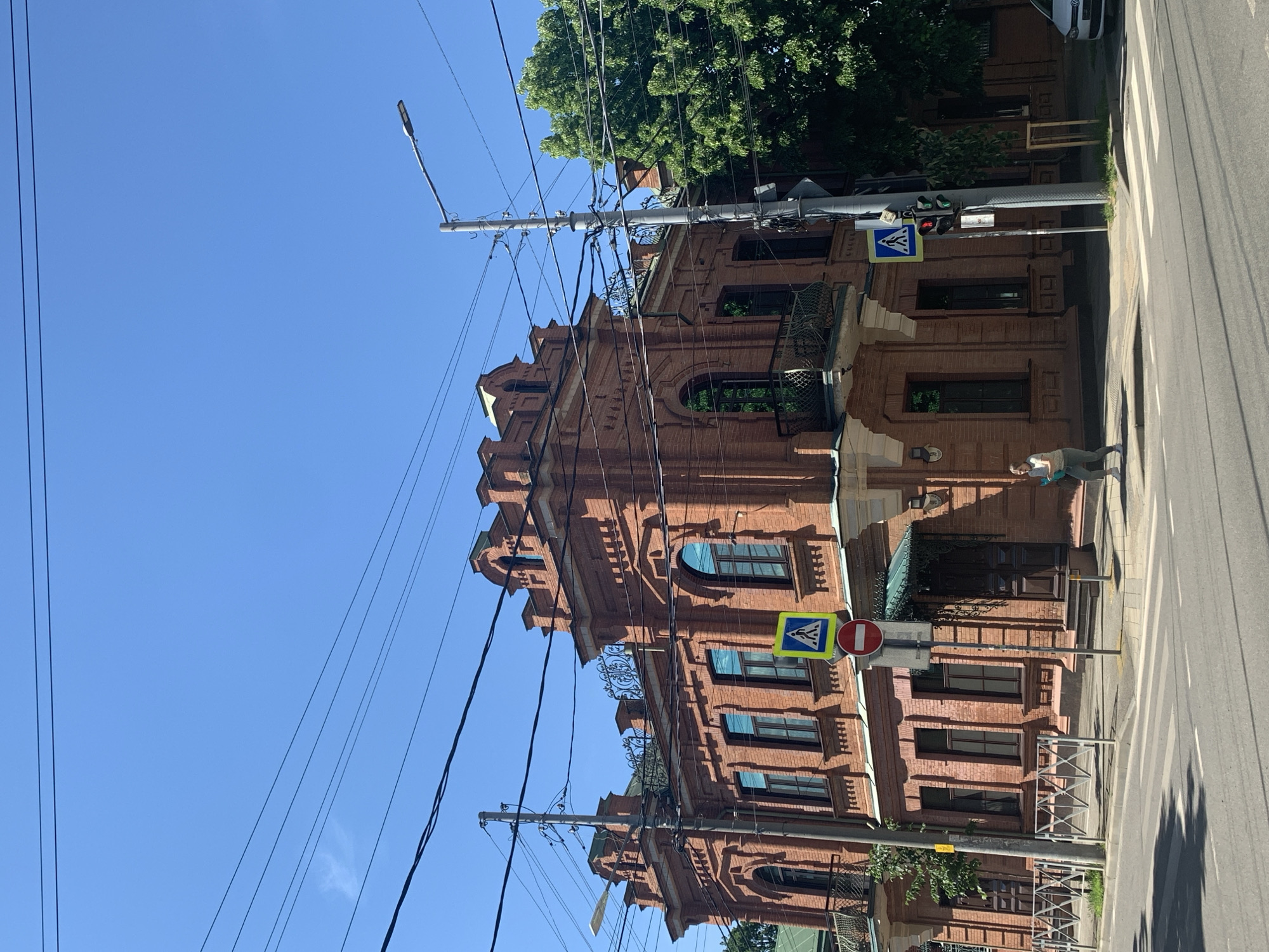 An urban street scene unfolds on a bright, clear day, showcasing a detailed, multi-story brick building on the right. The building features ornate architectural elements, including decorative cornices, arch-shaped windows, and balconies with wrought-iron railings. Numerous utility wires and power lines are strung across the foreground against a vivid blue sky.
On the left, a paved street with a pedestrian zebra crossing is visible. A person in light clothing, carrying a bag, walks on the sidewalk next to the crossing, moving away from the viewer. Street furniture includes a tall pole holding a modern street lamp and a traffic light which is red for vehicle traffic. Several road signs are present: a blue square sign with a white pedestrian symbol indicating a pedestrian crossing, and a red circular sign with a white horizontal bar, typically signifying "no entry." Metal barriers are positioned along the sidewalk and near the building's entrances. Lush green trees are visible above and behind the brick structure, and a white car is partially seen parked further down the street. Strong shadows cast by the sun suggest midday or early afternoon.
