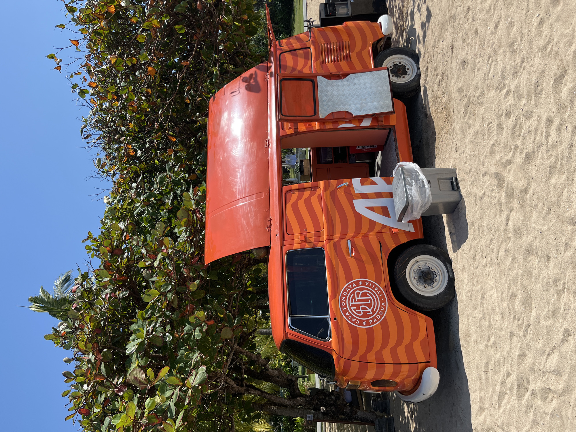 A bright orange vintage Volkswagen van, repurposed as a food truck, is parked on a sandy surface under a lush green tree. The van features wavy orange stripes and a logo on its side that reads "CASA FONDATA PADOVA ITALIA". The rear door and a top-opening panel are open, revealing the interior of the food truck. A grey trash can with a plastic liner stands beside the van. The background shows more foliage and a clear blue sky. The van is located on a beach in Kuta Selatan, Indonesia.