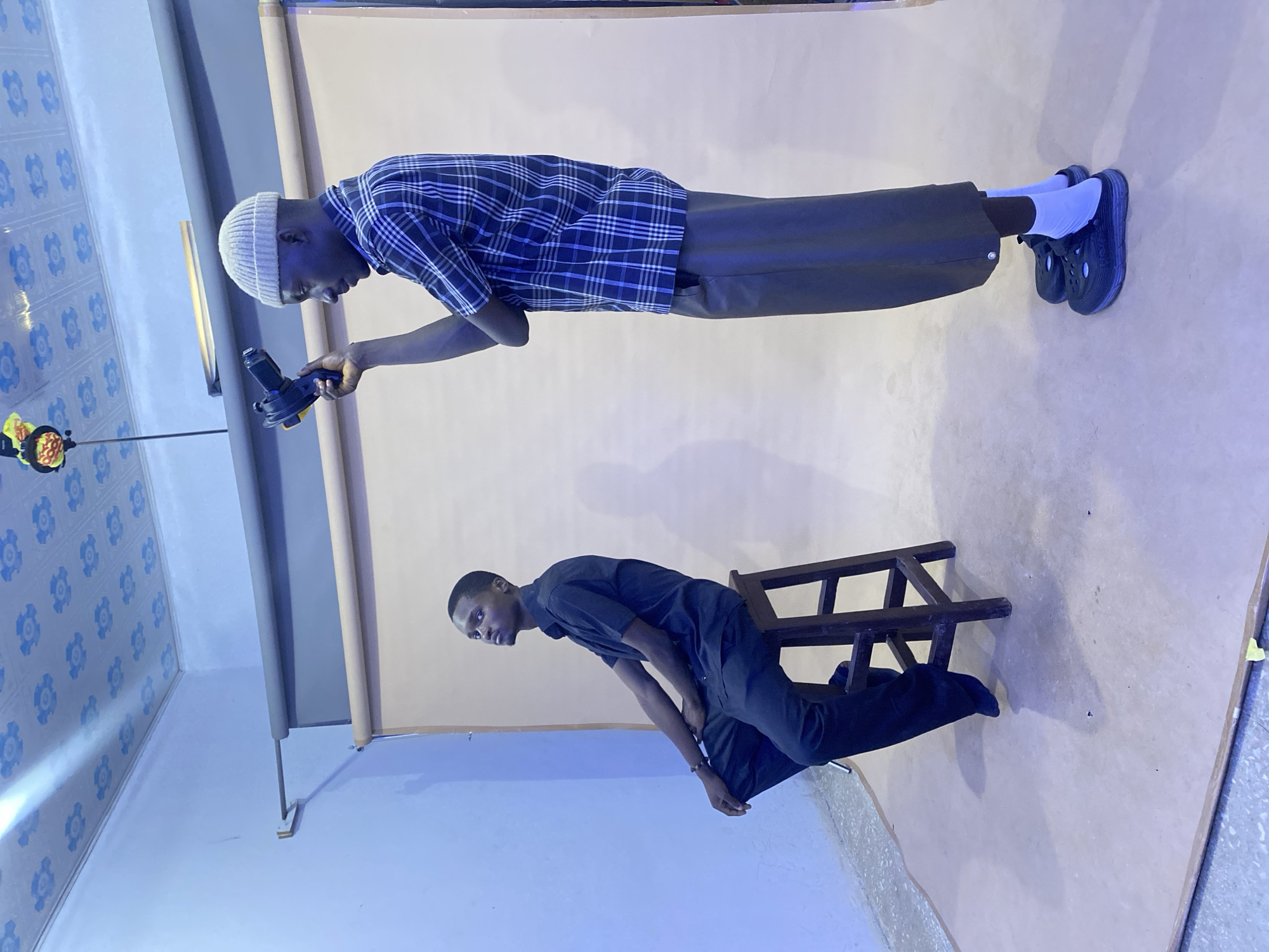 In a studio located in Gura Top, Nigeria, a photographer, wearing a light-colored beanie, a plaid shirt, and dark pants, stands to the right of the frame, holding a black camera with a yellow grip, aimed at a man seated on a wooden stool. The seated man, dressed in a black short-sleeved collared shirt and black pants, looks upwards and to his left. The studio features a light beige backdrop and a light blue wall on the left, with a patterned ceiling above. The floor is covered with a light brown material.