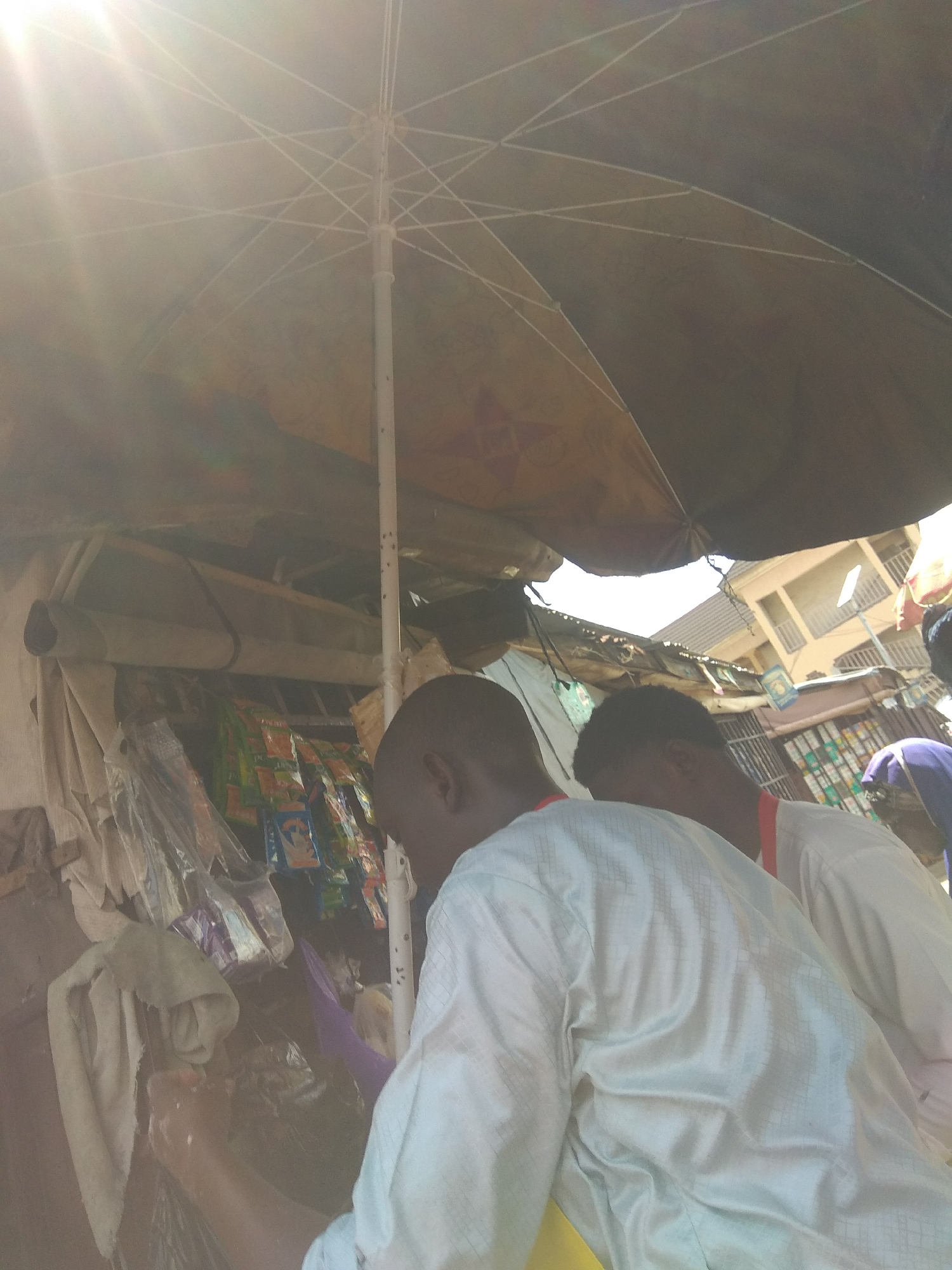 The image depicts a bustling street scene in Jalingo, Nigeria, under the harsh glare of the midday sun. A large, dark umbrella, its internal white spokes visible, provides shade over a makeshift stall laden with goods. The stall appears to be selling various packaged food items and possibly other small commodities, displayed in colorful wrappers. In the foreground, two individuals, likely customers or vendors, are engrossed in an interaction at the stall. One person, seen from the back and slightly to the left, is wearing a white traditional outfit with a checkered pattern. Their head is bowed as if examining or handling items. To the right, another individual, also in a white outfit, is partially visible, facing the same direction. Behind the stall, the architecture of Jalingo is visible, with buildings featuring balconies and multiple floors, suggesting a densely populated urban environment. The overall impression is one of everyday commerce and activity in a Nigerian city.