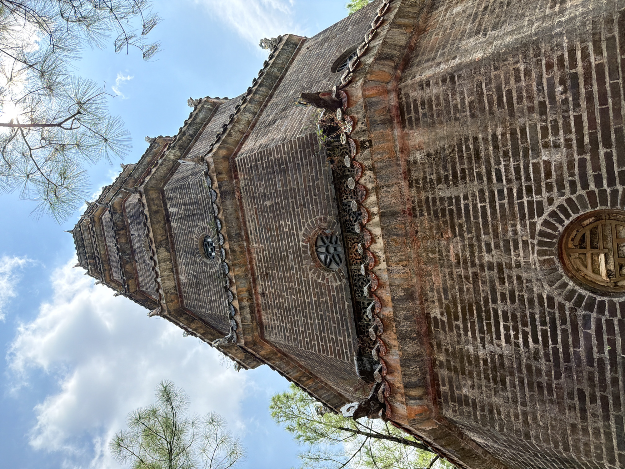 An upward-angle, medium shot captures the octagonal brick pagoda of the Tomb of Thieu Tri in Kim Long Ward, Vietnam, under a bright, partly cloudy blue sky. The pagoda, constructed from dark, weathered bricks, features multiple tiers with ornate, tiled eaves adorned with decorative dragon figures and roof ornamentation. Several circular, lattice-patterned windows are visible on different levels of the structure. Green trees with fine needles, likely pine, frame the top of the pagoda, with sunlight filtering through their branches. The lower portion of the pagoda exhibits significant discoloration and moss growth, indicating age and exposure to the elements. One circular window with a decorative wooden lattice is prominently featured on the lowest visible section of the pagoda. The overall scene is one of historical architecture set within a natural environment.