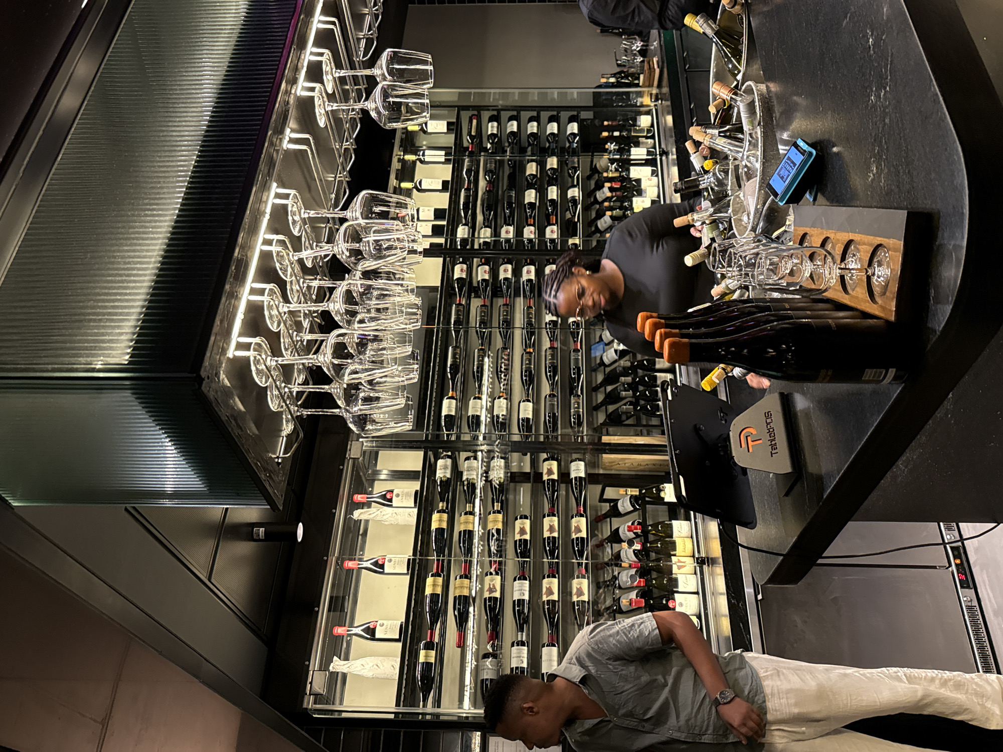 The scene depicts the interior of a wine bar, possibly located in Cape Town, South Africa, based on the subject matter. The focus is a glass-enclosed wine rack displaying numerous bottles of wine, predominantly red. Wine glasses hang upside down in racks, illuminated by a light source. A woman with braided hair and glasses is standing behind the counter, presumably attending to the wines. Another individual, a man with short hair, is seen looking at the lower portion of the wine rack. The bar top is a dark, speckled material, cluttered with wine bottles, glasses, and a POS system. A stainless steel refrigerator is visible.