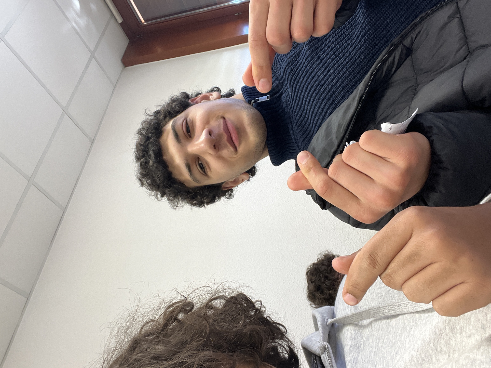 A low-angle shot shows two people indoors. The person in focus has curly dark hair, wears a dark blue zipped sweater, and a black puffy jacket. They smile at the camera and point both index fingers downwards. Partially visible in the foreground is the top of another person's head with curly brown hair, wearing a gray hoodie. The background features a white tiled wall and a window with a brown frame in the upper portion of the image. It's difficult to confirm the location in Schiltigheim, France, without additional contextual information.