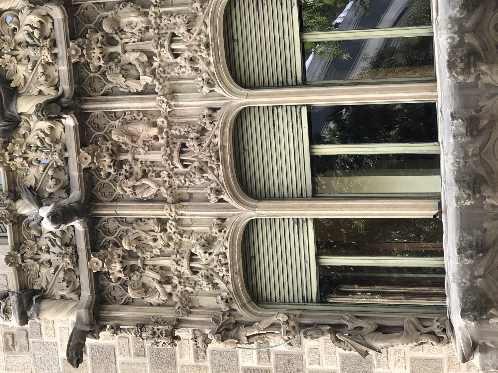 The image shows a close-up view of ornate stone carvings adorning the facade of a building. The carvings are intricate and detailed, featuring figures, foliage, and architectural elements. Three arched windows with green blinds are visible below the carvings. The building appears to be constructed of stone blocks. The lighting suggests it is daytime.