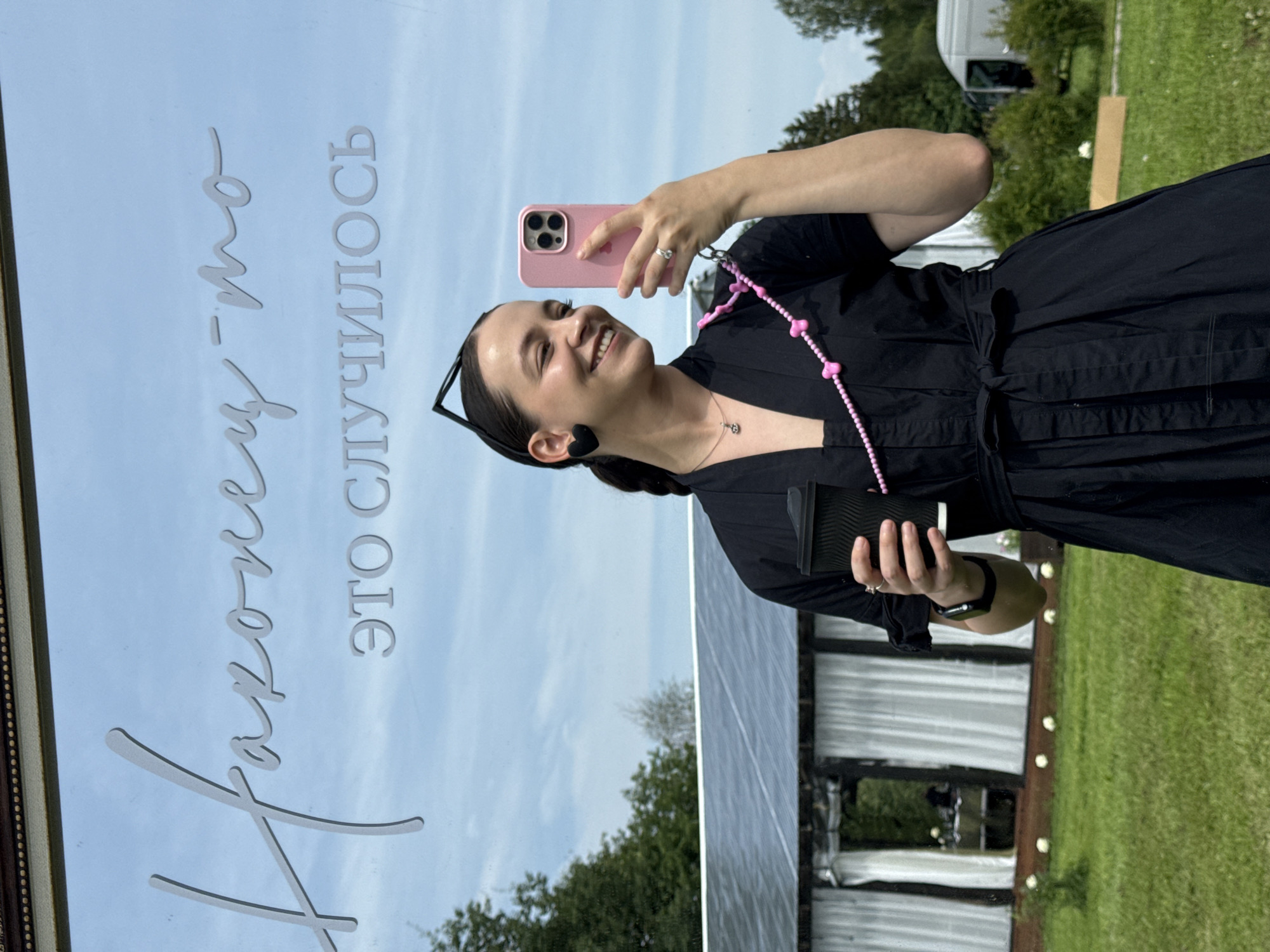 A woman is taking a selfie in front of a mirror, with the sky and trees reflected in it. The mirror has text written on it, "Наконец-то это случилось" (Finally, it happened). The woman is wearing a black dress and holding a black coffee cup in her left hand and a pink phone in her right hand. She is smiling and looking at her phone. There is a white tent with curtains in the background. The location is possibly the city of Svetloe, Russia, given the context of the provided crops. The scene is outdoors, with green grass and trees visible.