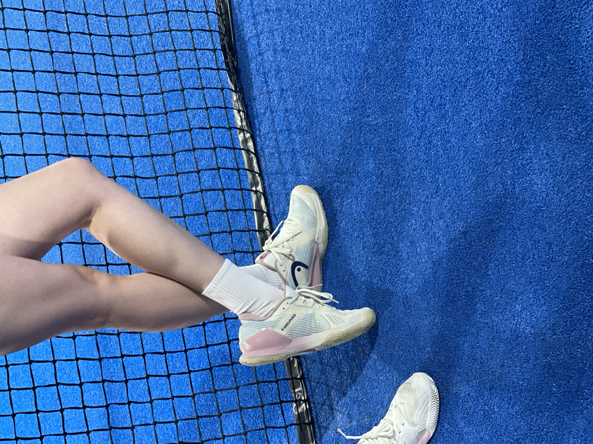 The image shows a close-up view of a person's legs and feet on a blue court, likely a tennis or padel court, with a black net in the background. The person is wearing white socks and white and pink athletic shoes. One shoe is the main focus, showing the "HEAD" logo and a blue accent with a swoosh design. The other shoe is partially visible on the left side of the frame. The court surface appears to be artificial turf. The lighting suggests an indoor or outdoor setting with diffused light. There is no indication of the location being in the city of Orenburg, Russia.
