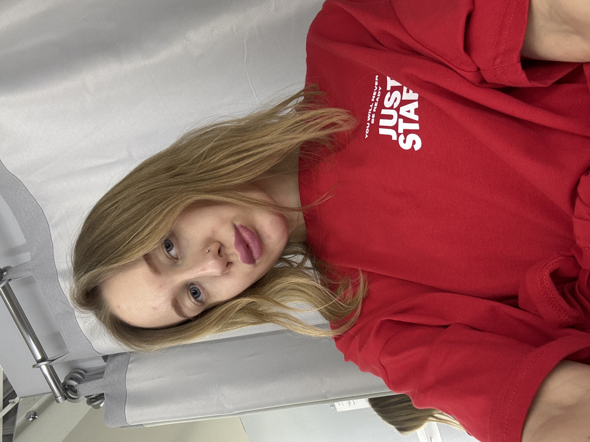 A young woman with blonde hair and blue eyes, wearing a red t-shirt with white lettering "JUST STAFF YOU WILL NEVER BE READY", is lying on a light grey medical bed. The photo was taken from a slightly angled, close-up perspective. A metal frame is visible on the left side of the bed. The setting appears to be a medical facility. The image does not give any information to place it in Rostov-on-Don, Russia.