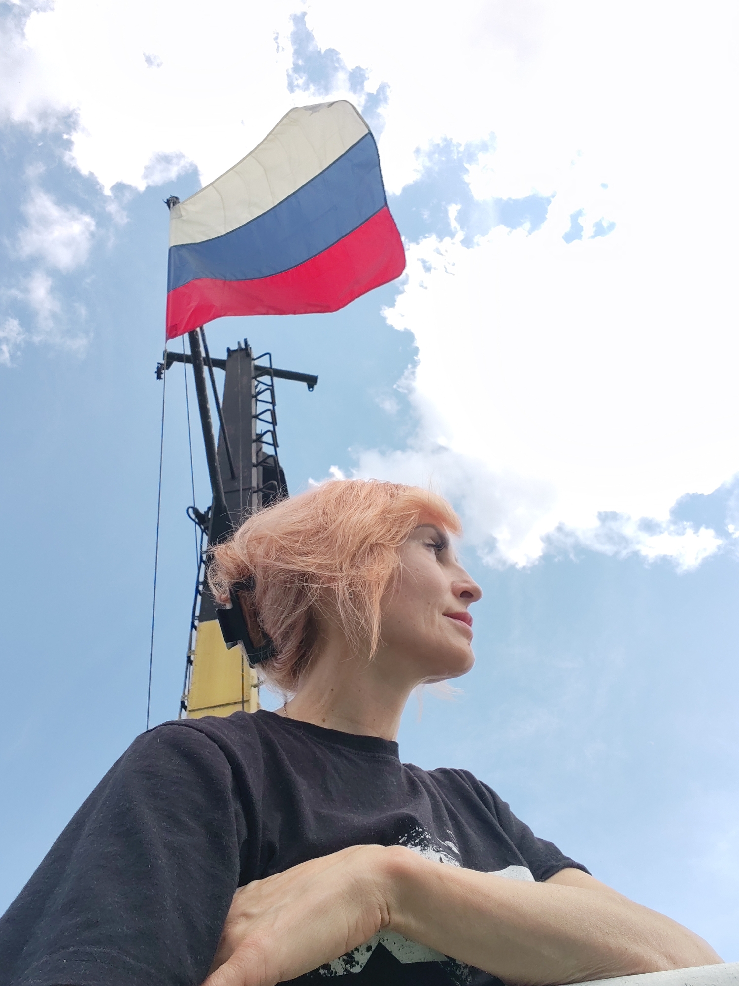 A low-angle, full-shot photograph captures a woman with short, pink-tinted hair and a black t-shirt, her arms crossed, as she looks upwards towards a Russian flag flying from a tall structure against a bright, cloudy sky. The woman is positioned in the foreground, with her profile visible as she gazes to the right. Behind her, the Russian flag, featuring white, blue, and red horizontal stripes, is prominently displayed. The flag is attached to a mast or a similar upright structure, which appears to be part of a building or installation. The background consists of a clear blue sky dotted with white clouds. The overall environment suggests an outdoor setting, possibly in the city of Belousovo, Russia, given the presence of the national flag.