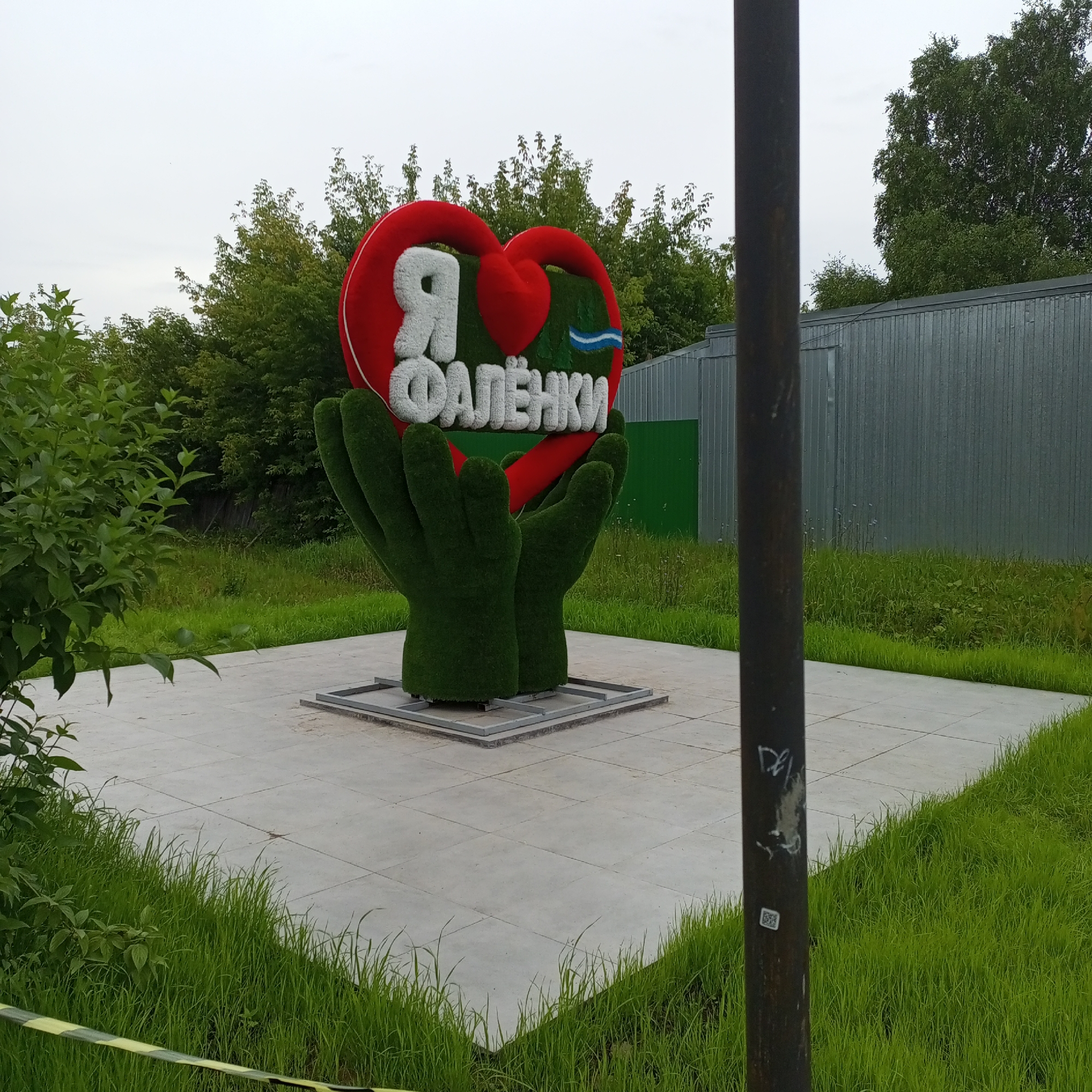 The image shows a monument in the city of Falenki, Russia, dedicated to the city itself. The monument features a large, stylized red heart atop two green, hand-like sculptures made of artificial turf. Inside the heart, the white Russian letters "Я" and "ФАЛЁНКИ" are prominently displayed, with "Я" meaning "I" and "ФАЛЁНКИ" being the name of the city. A small blue and white wavy line is positioned between the two words. The monument is situated on a square paved area, surrounded by green grass and trees. In the background, there is a grey corrugated metal fence and more trees. A dark, weathered metal pole with some graffiti is visible in the foreground, partially obscuring the view of the monument. The sky is overcast, suggesting a cloudy day. The overall scene depicts a civic monument celebrating local identity in the urban environment of Falenki.