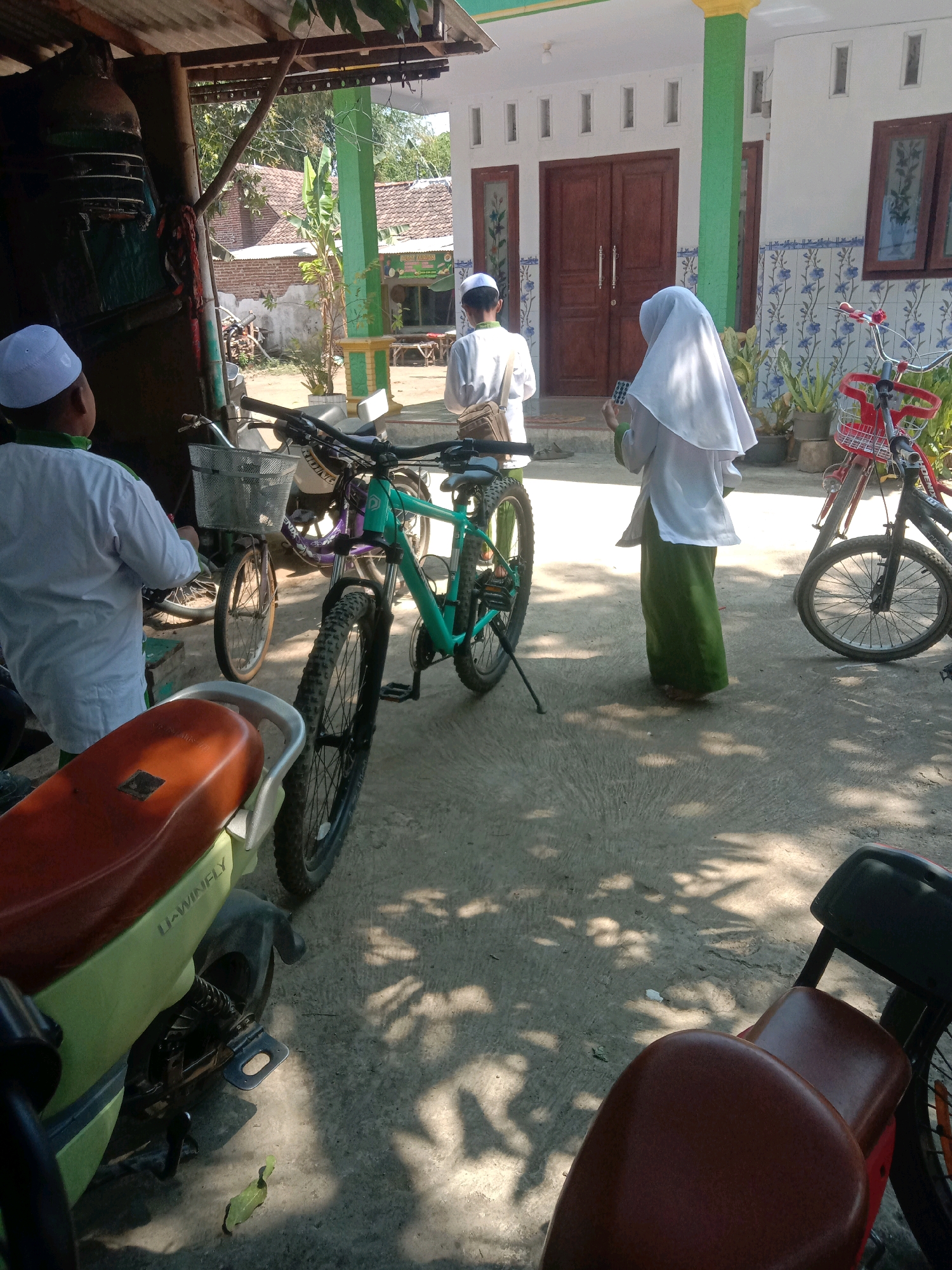 The image captures a sunny day in front of a building in Ngempit, Indonesia. A young boy in a white cap and traditional Islamic attire stands facing away from the camera, his left hand holding a mobile phone. Beside him, a person wearing a white hijab and a long green skirt is also seen from behind, holding a similar mobile phone. They appear to be waiting or observing something. Several bicycles are parked around them, including a bright turquoise mountain bike in the foreground. A light green motorcycle with a brown seat is also visible. The building in the background features a light-colored facade with blue and white floral patterned tiles and large wooden doors. The environment suggests a residential area or a religious institution, possibly a school, given the attire of the individuals.
