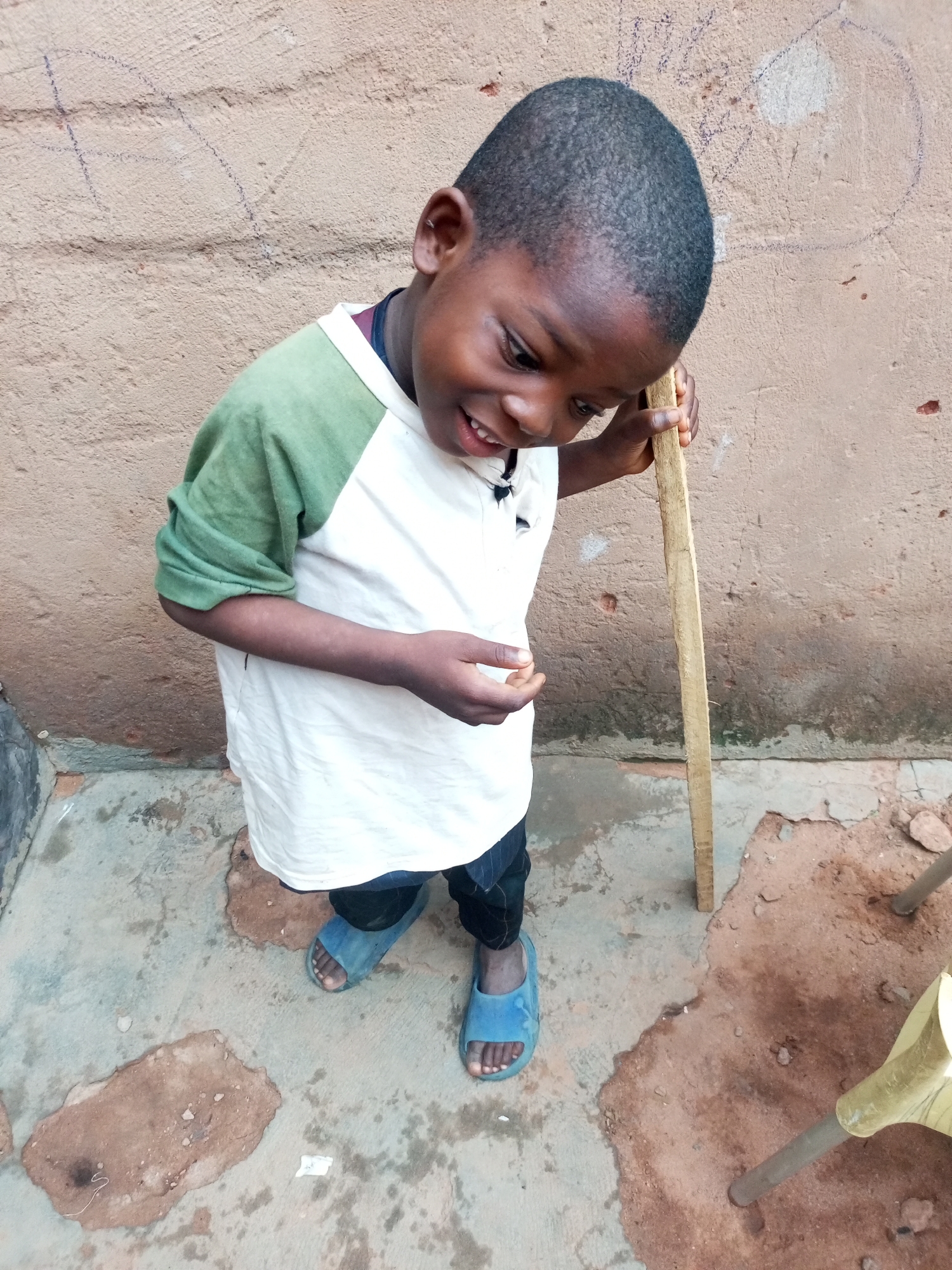 A young Black boy, appearing to be around five years old, stands outdoors next to a rough, tan-colored wall, possibly made of stucco or cement. He is wearing a short-sleeved, raglan t-shirt with green sleeves and a white body. He has dark, short hair and is looking down and to his right, with a slight smile. His right hand is resting on a light-colored wooden stick, held vertically. He is also wearing dark pants and blue slide sandals. The ground beneath him is a mixture of concrete and packed dirt, with some scattered debris. A yellow plastic chair leg is visible to the right. The overall scene appears to be in an urban or semi-urban environment in Tempure, Nigeria.