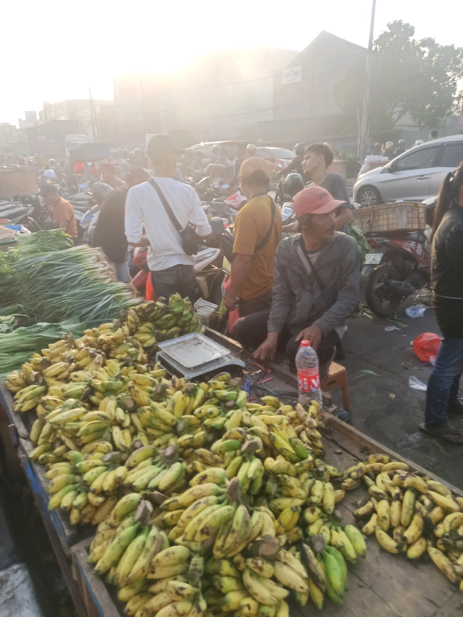 This is a wide-angle, medium-shot photograph captured from a slightly elevated perspective, looking down upon a bustling outdoor market in Cikarang, Indonesia. The scene is awash in the bright, hazy light of early morning, with the sun low in the sky, creating a slightly overexposed and diffused atmosphere.
The foreground is dominated by two large wooden stalls overflowing with bananas. The stall on the left displays bunches of bananas that are predominantly green with hints of yellow, while the stall on the right features more mature, yellow bananas with some brown spots, indicating ripeness. Interspersed with the bananas on the right stall is a metal weighing scale.
Several people are visible amidst the market activity. A man in a red cap and a grey jacket is seated on a small stool, his hands resting on his lap. Beside him is a plastic bottle of water. To his left, another man in a yellow shirt stands, looking away from the camera. In the background, a man in a white shirt with a dark bag slung across his shoulder is walking away. Other individuals, blurred by the distance and the haze, are scattered throughout the market.
The market appears to be a vibrant hub of commerce. Beyond the banana stalls, there are mounds of green vegetables, possibly scallions or leeks, piled high. Various motorcycles are parked and in motion, suggesting transportation and deliveries. In the distance, several cars, including a silver sedan, are visible on a paved road. The background is characterized by buildings and structures typical of an urban environment, shrouded in the morning mist. The overall impression is one of a lively and active marketplace at the start of the day.