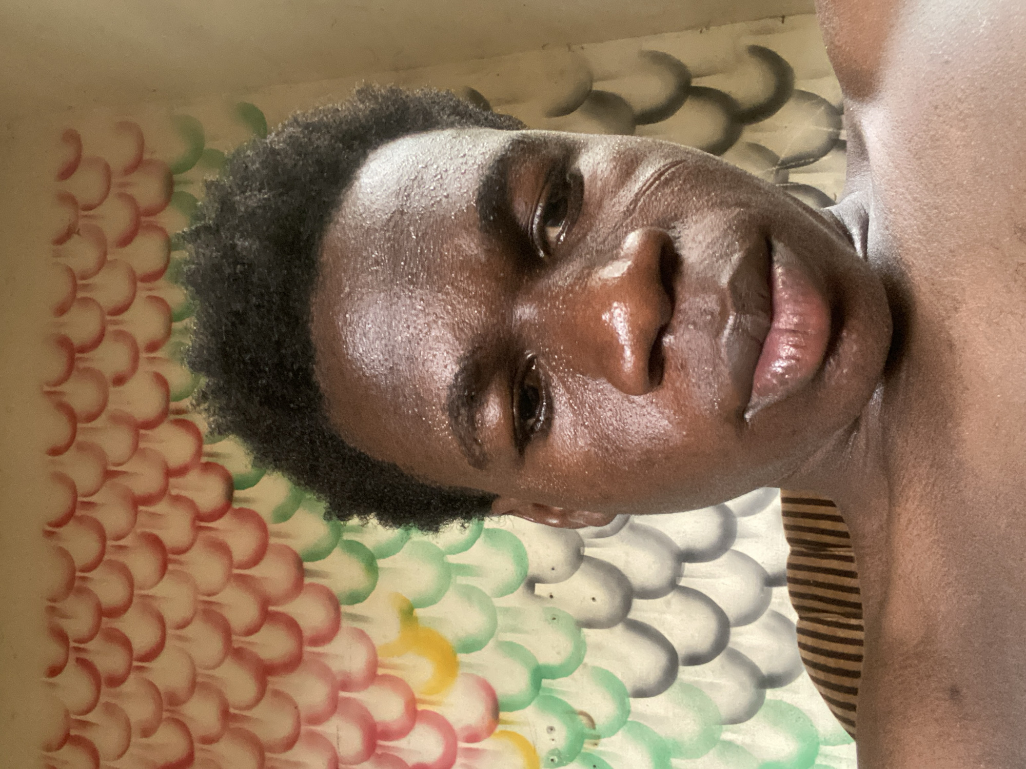 A close-up portrait features a young Black individual, likely male, with short, dark, kinky hair, presented in a slightly low-angle perspective. Their face and upper body are visibly glistening, suggesting sweat or moisture on their dark complexion. The individual looks directly at the camera with a neutral expression.
The background is a wall adorned with a distinct pattern of overlapping, semi-circular, or scale-like shapes. These shapes are arranged in horizontal bands of color: beginning with light red and pink on the far left, transitioning to light green in the middle, and fading into shades of grey and black towards the right side of the frame, behind the subject's head. A subtle hint of yellow shapes can also be seen below the green section. The individual appears shirtless, though a small section of a striped brown and black garment is visible on their lower right side.
The lighting is bright, creating prominent highlights on the person's skin. The scene suggests an indoor or shaded outdoor setting. No specific activity is taking place, and there is no visible text in the image. This moment is captured in Mararaba, Nigeria.