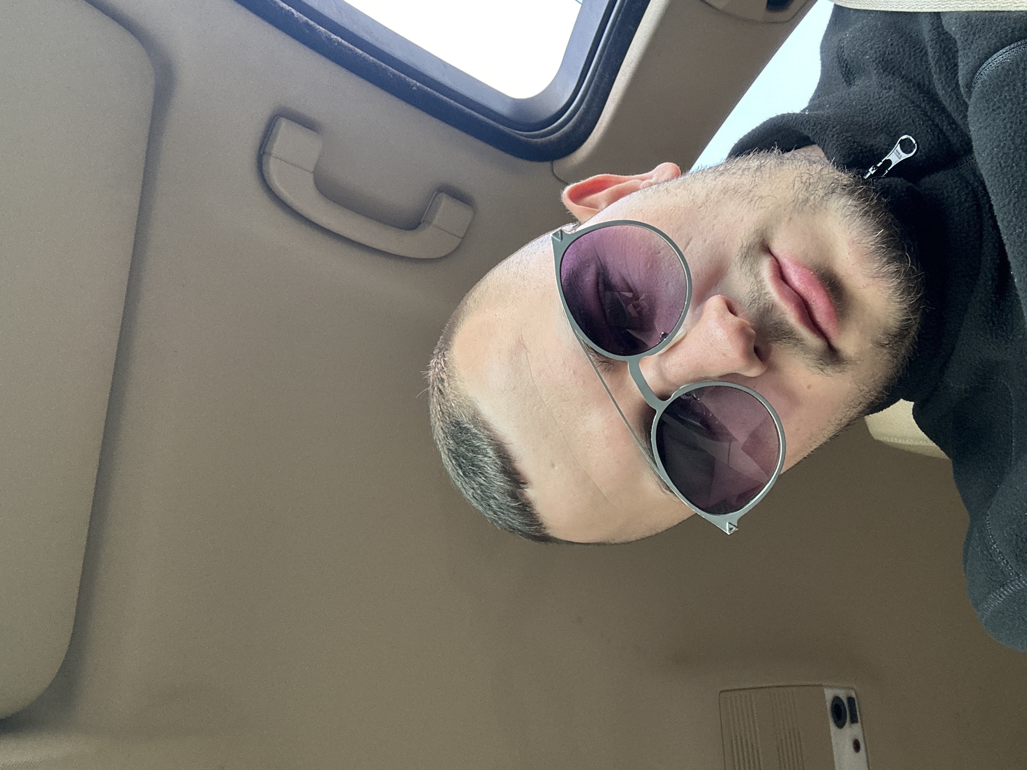 A man is inside a car in Plovdiv, Bulgaria. He is wearing a black jacket and sunglasses with round, purple-tinted lenses. The interior of the car is beige. There's a handle on the car's ceiling near the sunroof. A phone is partially visible near the bottom of the frame. The man's head is tilted, and the view is from below.