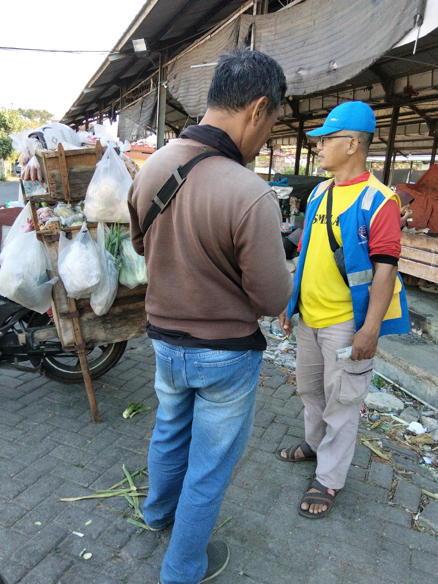 Two men are standing on a cobblestone street in front of a market stall in Banyudono, Indonesia. The man on the left is wearing a brown long-sleeved shirt and blue jeans. He has his back to the camera and is looking at the man on the right. The man on the right is wearing a yellow shirt with a blue and yellow vest and a blue baseball cap. He is wearing khaki pants and sandals. He is holding a bill in his hand. In the background, there is a market stall with various items displayed, including bags of produce. The street is lined with debris.