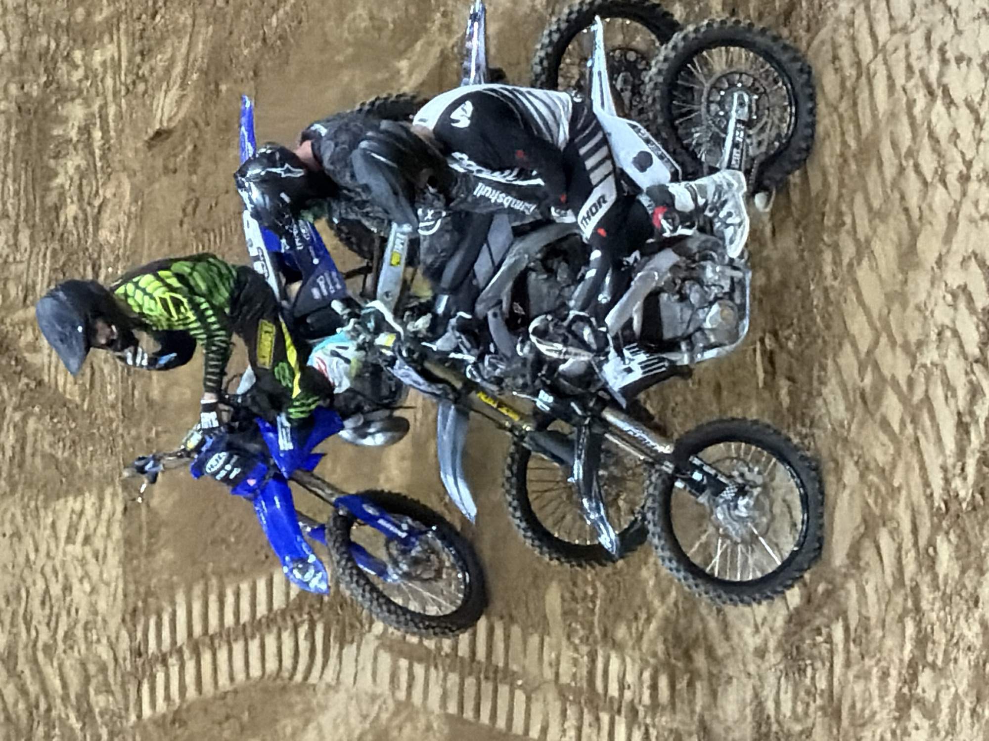 This image shows a motocross race with three riders on dirt bikes on a dirt track. The riders are wearing helmets and protective gear. The track appears to be in a stadium or arena setting, with spectators in the background, suggesting a professional event. The terrain is made up of compacted dirt with distinct tire tracks. The bikes are diverse in color, with one blue and white, one black and white, and another primarily black. The image captures a moment of intense competition, with riders positioned closely together. The lighting suggests an indoor event with artificial lighting. The specific location in Sacramento, USA, cannot be determined from the image alone.