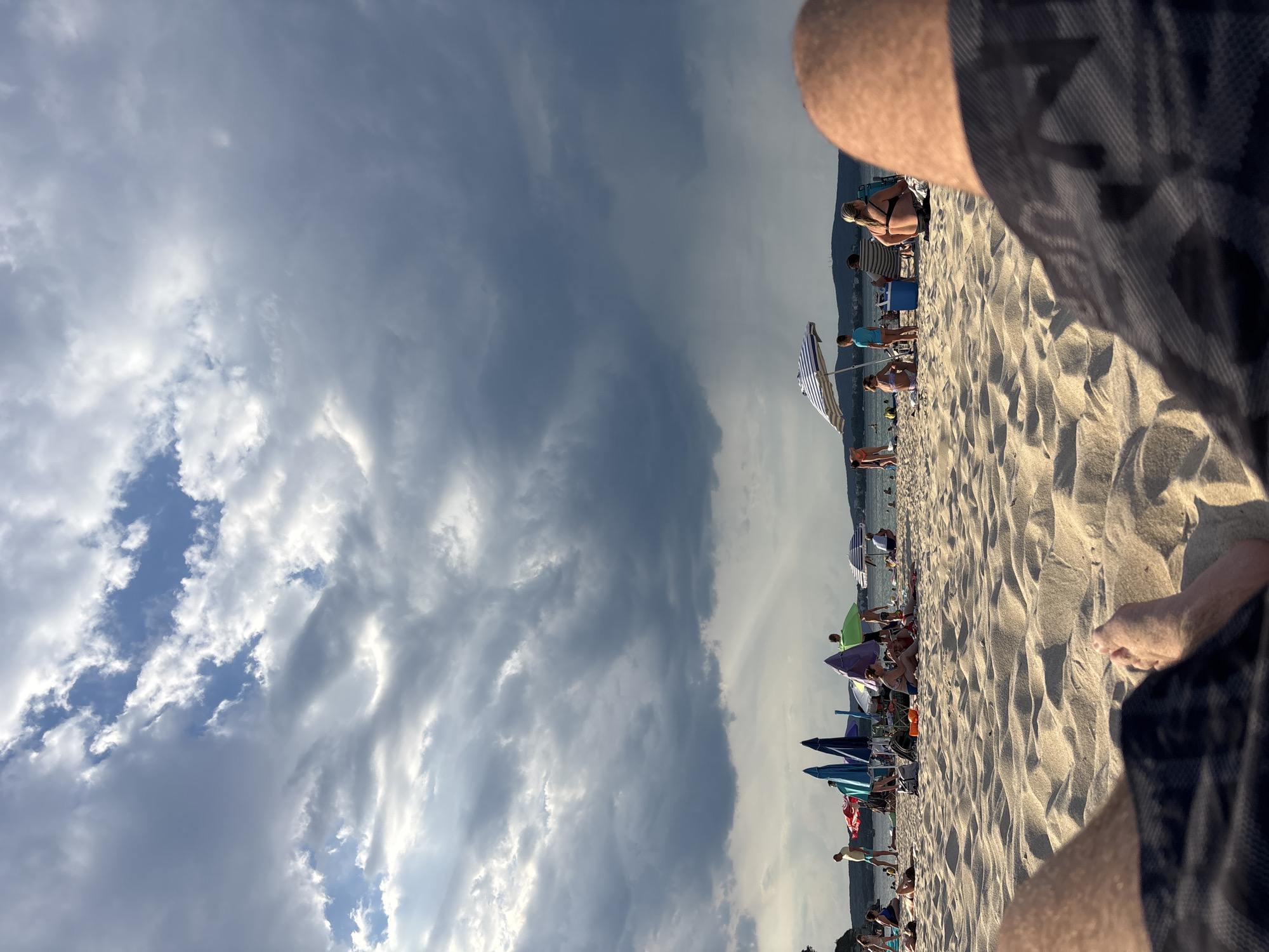 The image depicts a beach scene in Kiten, Bulgaria, under a dramatic sky. The foreground is dominated by the legs of the photographer, reclining on the sand. The beach itself is a expanse of light-colored sand, with ripples and dunes created by the wind. Scattered across the middle ground are various beachgoers and their belongings. Several colorful beach umbrellas provide shade, with people sitting or standing near them. Some individuals are seen in swimsuits, engaged in typical beach activities like sunbathing or socializing. The middle distance shows the water of the Black Sea, with people in the water and along the shoreline. In the background, rolling hills and a distant coastline are visible. The sky is a focal point, filled with heavy, dark storm clouds that are breaking to reveal patches of lighter sky and hints of sunlight, creating a striking contrast. The overall atmosphere suggests an approaching or departing storm, adding a sense of drama to the otherwise leisurely beach setting.