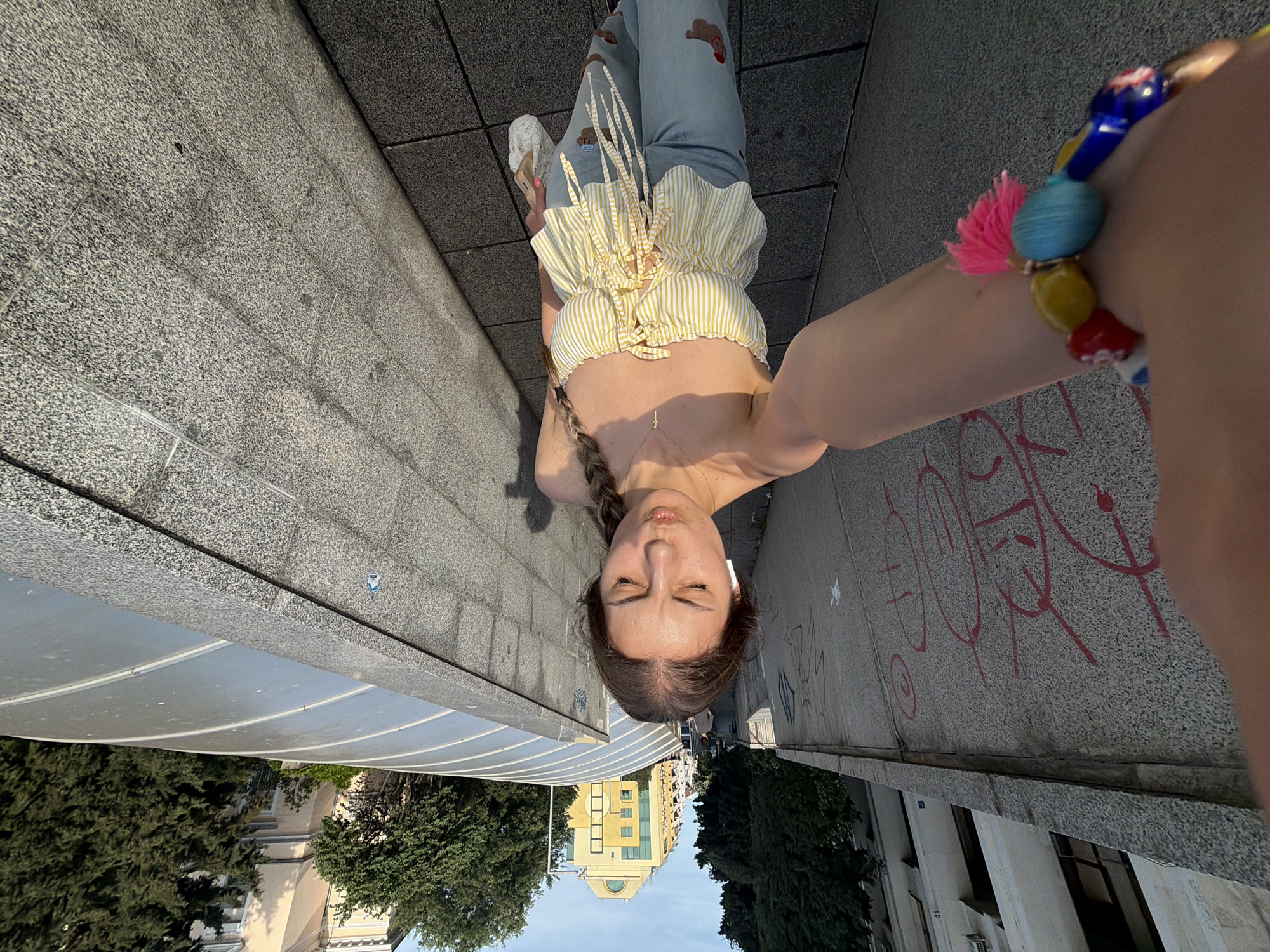 This media file presents a self-portrait of a young woman, captured from a low angle. The original image is rotated 90 degrees counter-clockwise; this description will orient the scene correctly, as if the woman is standing and looking upwards. She is the central subject, with her eyes closed and a serene, relaxed expression. Her light brown hair is styled in a braid, falling over her left shoulder. She wears a cheerful yellow and white striped crop top with ruffled details and multiple ties at the front. Her light blue jeans, visible below, feature distressed areas and distinctive brown teddy bear-shaped patches. A delicate gold-colored necklace with a small cross pendant adorns her neck, and small white earbuds are visible in her ears. On her right wrist, which is extended to hold the camera for the selfie, she wears a colorful beaded bracelet accented with a vibrant pink tassel.
The setting is an urban environment in Burgas, Bulgaria. The ground beneath her is paved with grey, speckled square tiles. To her left, a darker, rougher concrete or stone surface, possibly a curb or low wall, runs parallel. To her right, another similar grey surface is visible, covered in red graffiti consisting of abstract shapes and lines.
In the background, under a clear blue sky, stand tall buildings and lush green trees. On the left side, a light-colored building, possibly yellow or beige, with multiple windows and balconies, is visible. A curved, translucent or reflective structure, possibly a modern shelter, stretches above this area. On the right, more buildings and dense green foliage contribute to the urban landscape. The warm, bright sunlight suggests it is late afternoon or early evening, casting soft shadows. The overall mood is peaceful and candid, capturing a personal moment in a bustling city environment. No clear, legible text is visible, only stylized red graffiti marks.