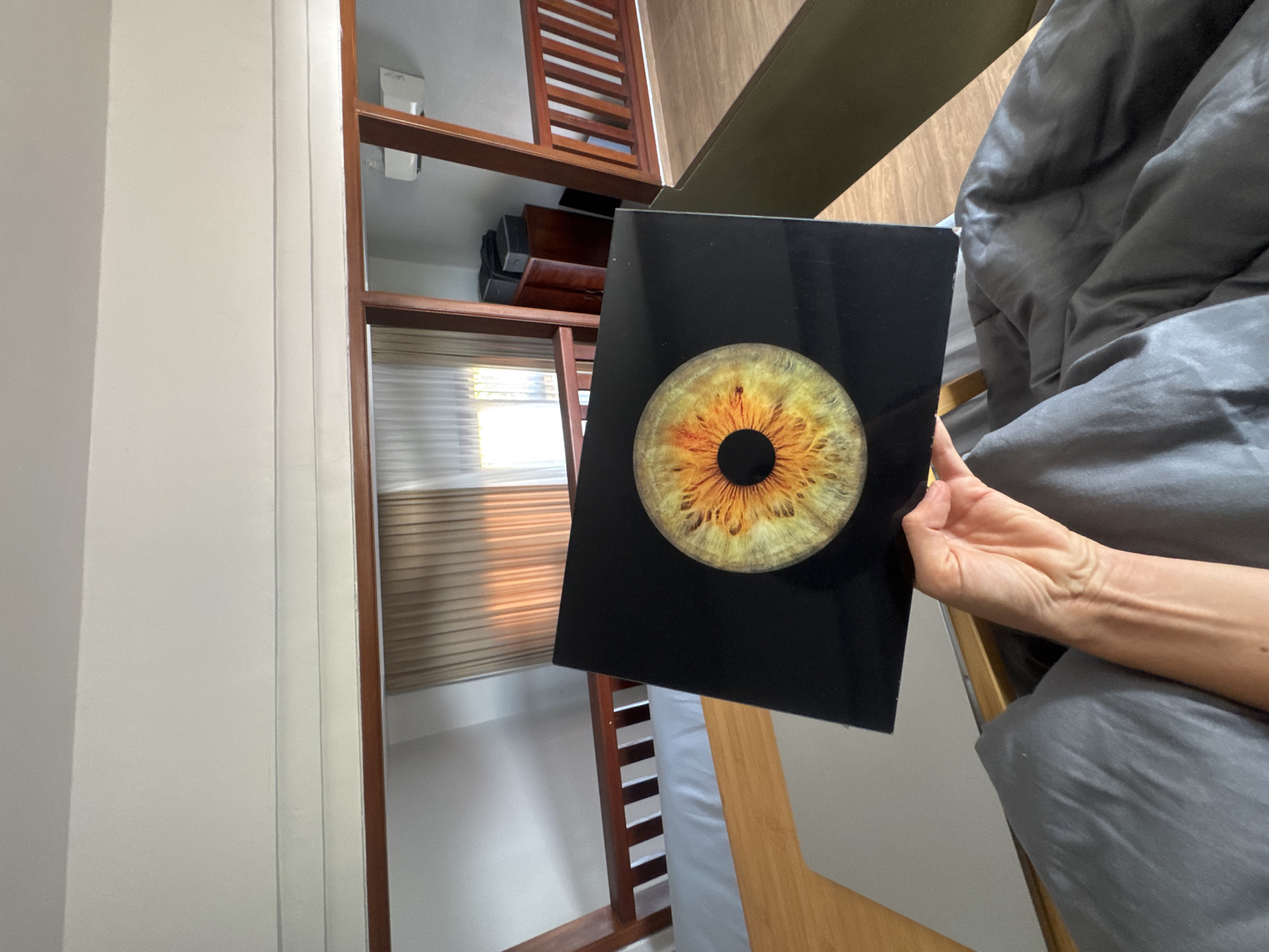 The image depicts a person holding a rectangular black object with a close-up image of a human iris on it. The iris is predominantly yellow and orange with some green and brown tones. The person's arm and hand are visible, holding the object over a bed with gray bedding. In the background, a wooden structure, possibly a bunk bed or a loft, is visible, along with a window with light-colored curtains. The environment appears to be an indoor setting, possibly a bedroom. The image does not provide any specific information about the location in North Kuta, Indonesia.