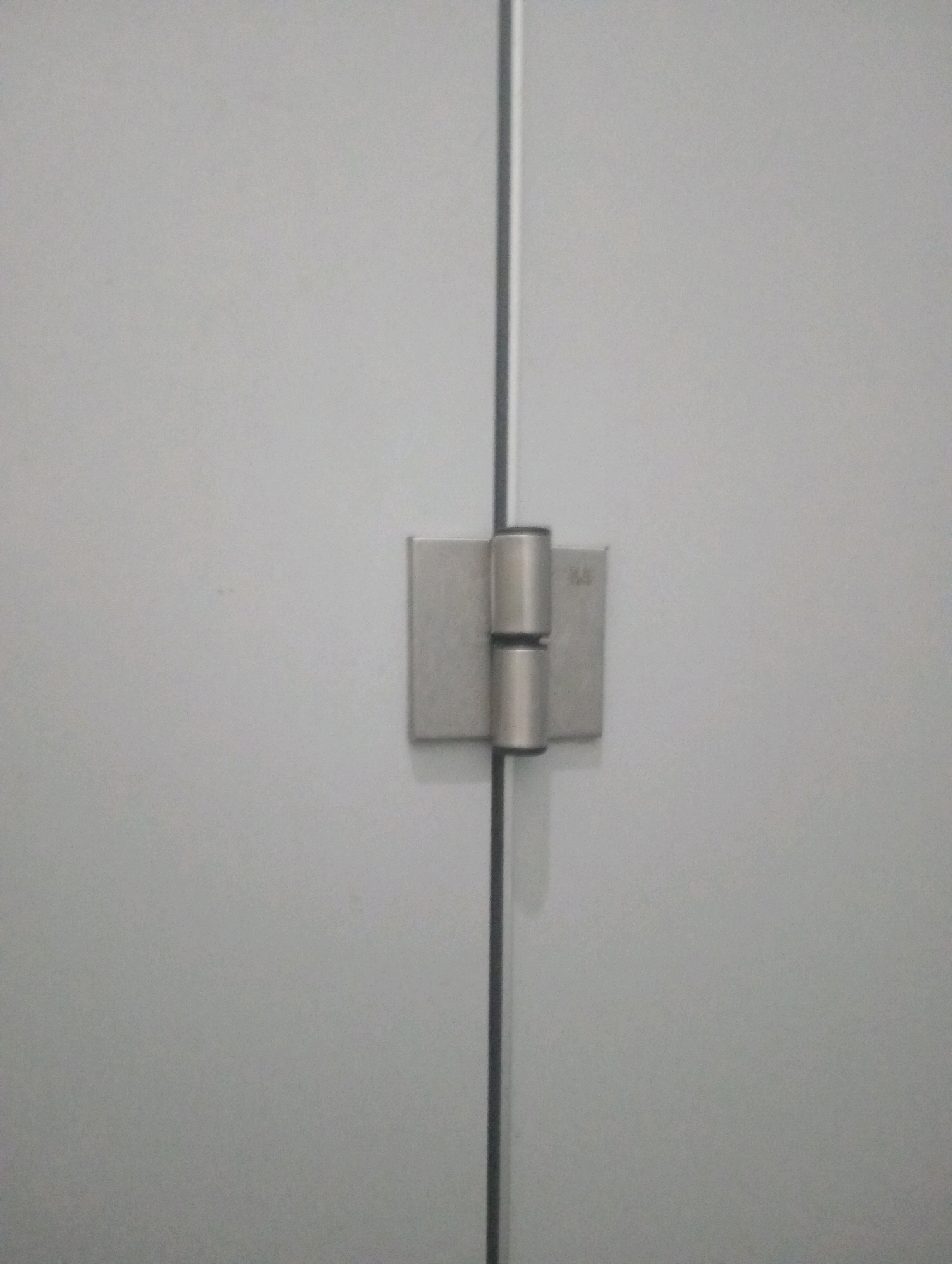 A close-up, eye-level, medium shot captures a stainless steel hinge connecting two plain, light gray surfaces that meet vertically down the center of the frame. The hinge is centrally located and features two cylindrical barrels joined together. The surfaces appear to be a door or wall panel. The background is a uniform light gray with subtle speckling, suggesting a matte finish. No people or actions are depicted. The image is devoid of any environmental context that would place it in Batu, Indonesia.