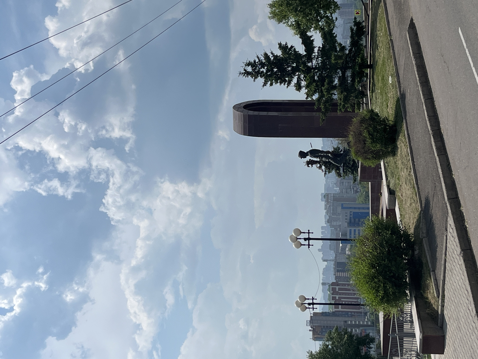 The image depicts a view from a city overlook in Krasnoyarsk, Russia, on a cloudy day. In the foreground, there is a paved walkway and a road. Behind the walkway, there are manicured bushes and grassy areas. Prominently featured is a monument consisting of a large, dark arched structure and a statue of a figure standing next to it. Several streetlights with multiple globes are visible, lining the walkway. In the background, a cityscape of buildings stretches across the horizon under a sky filled with white and gray clouds. Power lines are strung across the upper portion of the frame.