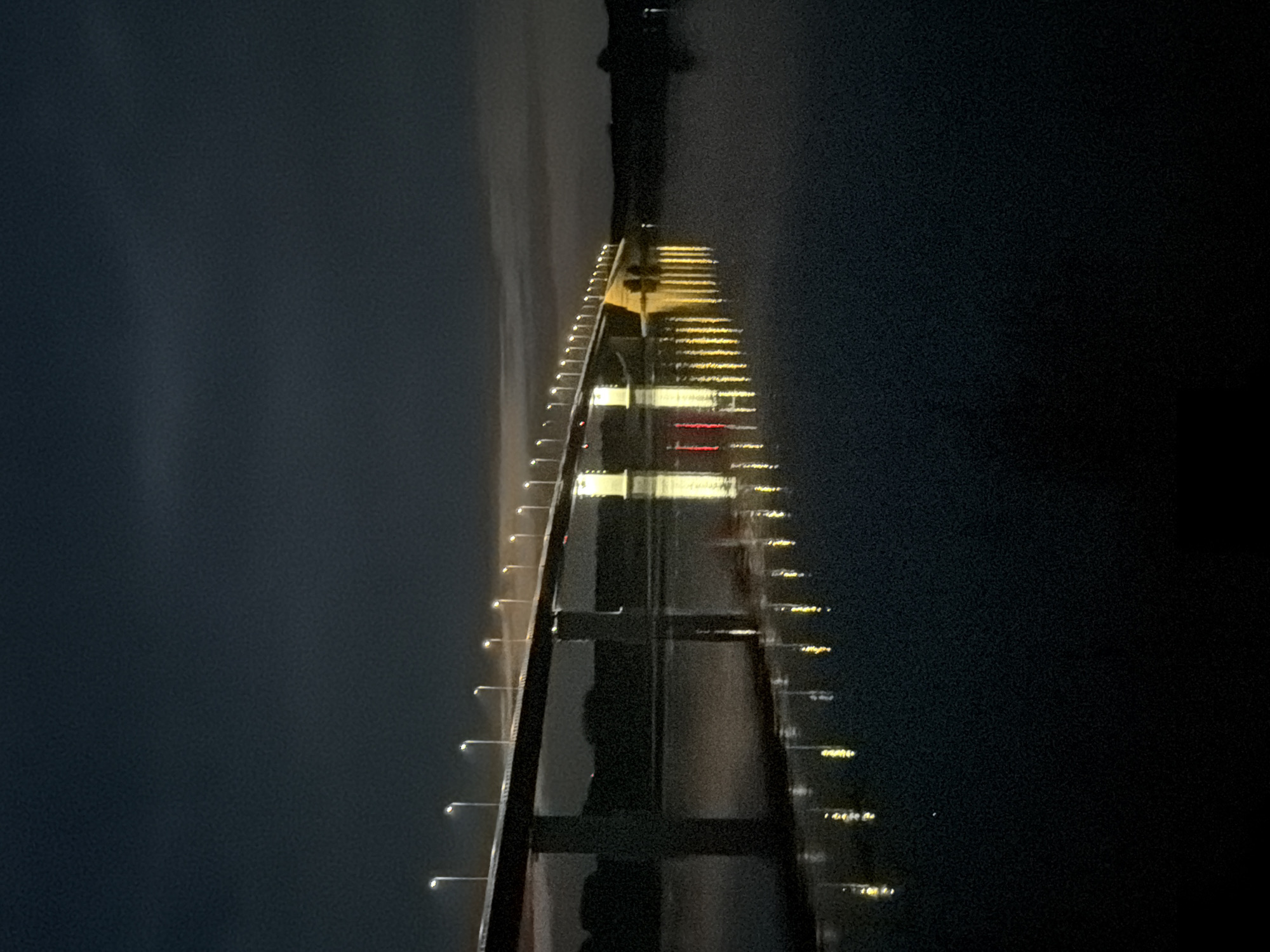 The image depicts a long bridge illuminated by streetlights at night. The bridge spans across a body of water, likely a river or lake, with the lights reflecting on the dark surface. The sky is dark and cloudy, suggesting nighttime. The bridge is supported by concrete piers. The scene appears to be located in Tarasovo, Russia, although specific landmarks are not visible to confirm this. The image is a static shot with no visible people or activity.