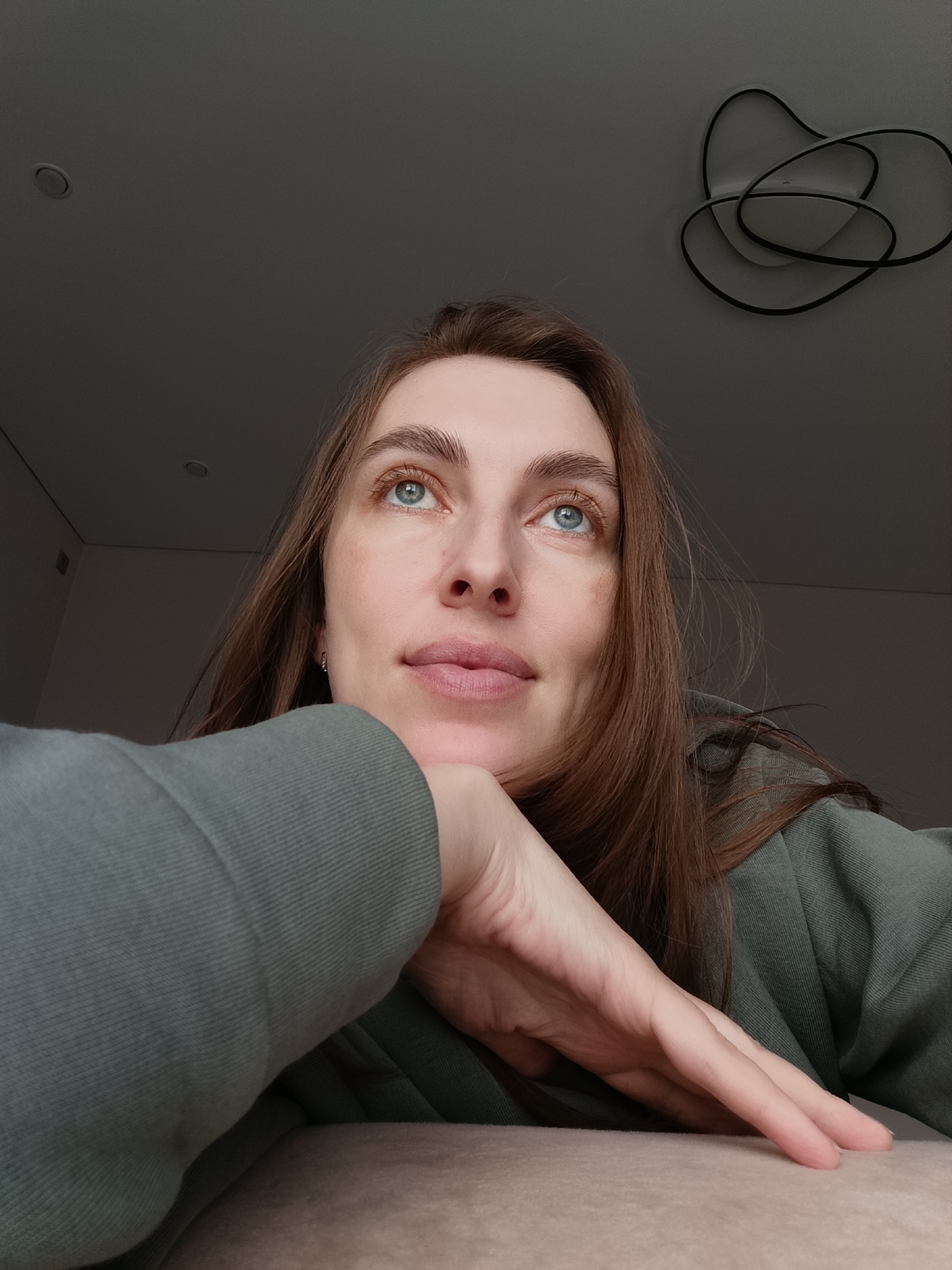 A woman with long brown hair and light blue eyes is looking upwards in an indoor setting. She is wearing a green sweatshirt and her left hand is resting under her chin. The background is a grey wall with a modern-looking ceiling light fixture. There is also a recessed light visible in the upper left of the frame. The location cannot be determined to be Chaikovsky, Russia from this image.