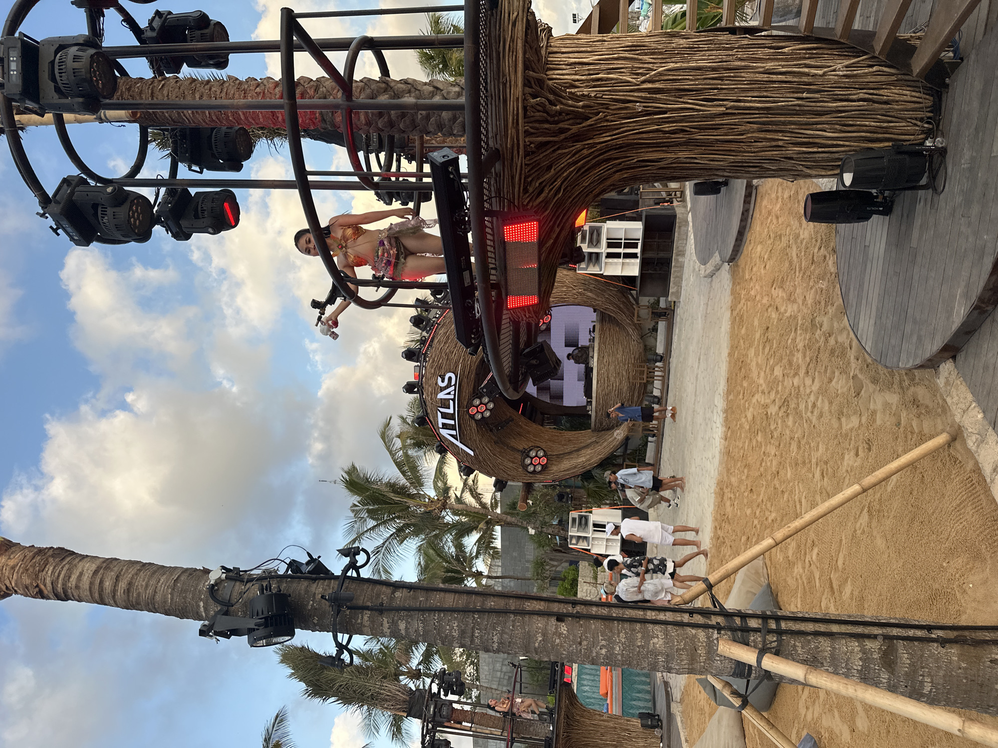 This image shows the exterior of what appears to be a beach club or outdoor event venue in North Kuta, Indonesia. The venue has a large, crescent-shaped architectural feature made of woven or thatched material with the word "ATLAS" displayed prominently. There is a stage area with sound equipment and lighting. A woman in swimwear is perched on a metal structure, possibly performing or serving drinks. People are visible in the venue, seemingly enjoying the event. The environment includes palm trees and sand, typical of a beach setting. The weather appears sunny with some clouds in the sky.