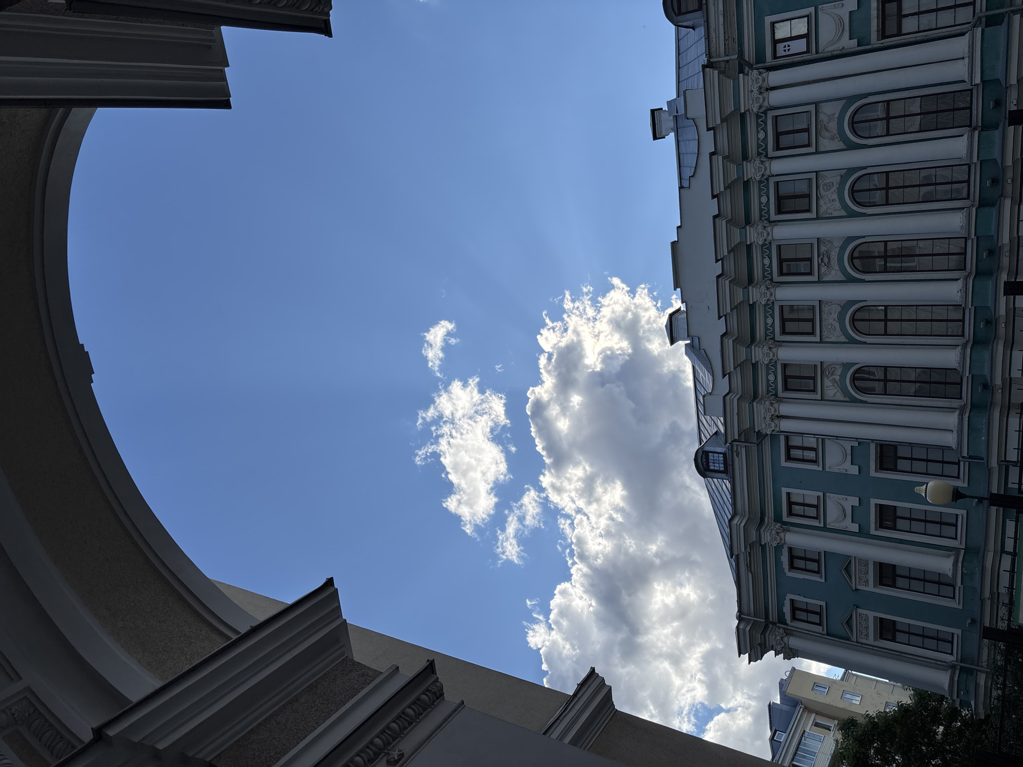 The image captures a view of a building with light blue facade and ornate white columns against a bright blue sky with scattered white clouds. The building has a symmetrical design with multiple windows, some arched and some rectangular. The roof is dark and sloped, with chimneys visible. In the foreground, part of an arched structure frames the scene, likely a part of a larger building or an architectural element. The lighting suggests it is daytime, with the sun casting shadows. The scene appears to be an urban environment, focusing on architectural details and the sky. Based on the provided location, this is a view of a building in Novaya Usman', Russia.