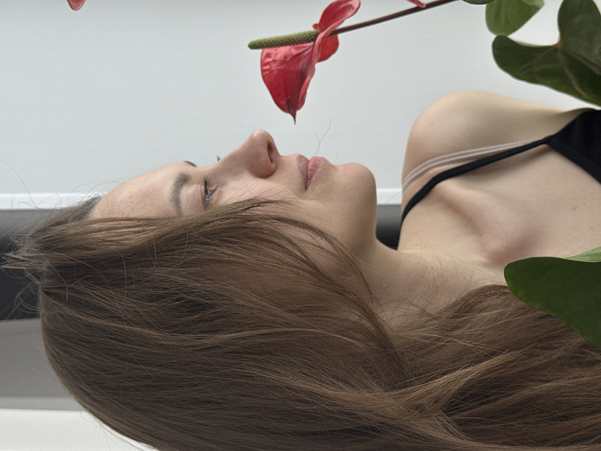 A young woman with brown hair is lying down, looking upwards towards a red anthurium flower. The flower is held above her face, with its spadix pointing down in her general direction. She has light skin and is wearing a black top with thin straps. Green leaves are partially visible, likely belonging to the same plant as the flower. The background appears to be a white or light-colored surface. The image was not taken in the city of Chaikovsky, Russia, as I have no information to suggest that it was.