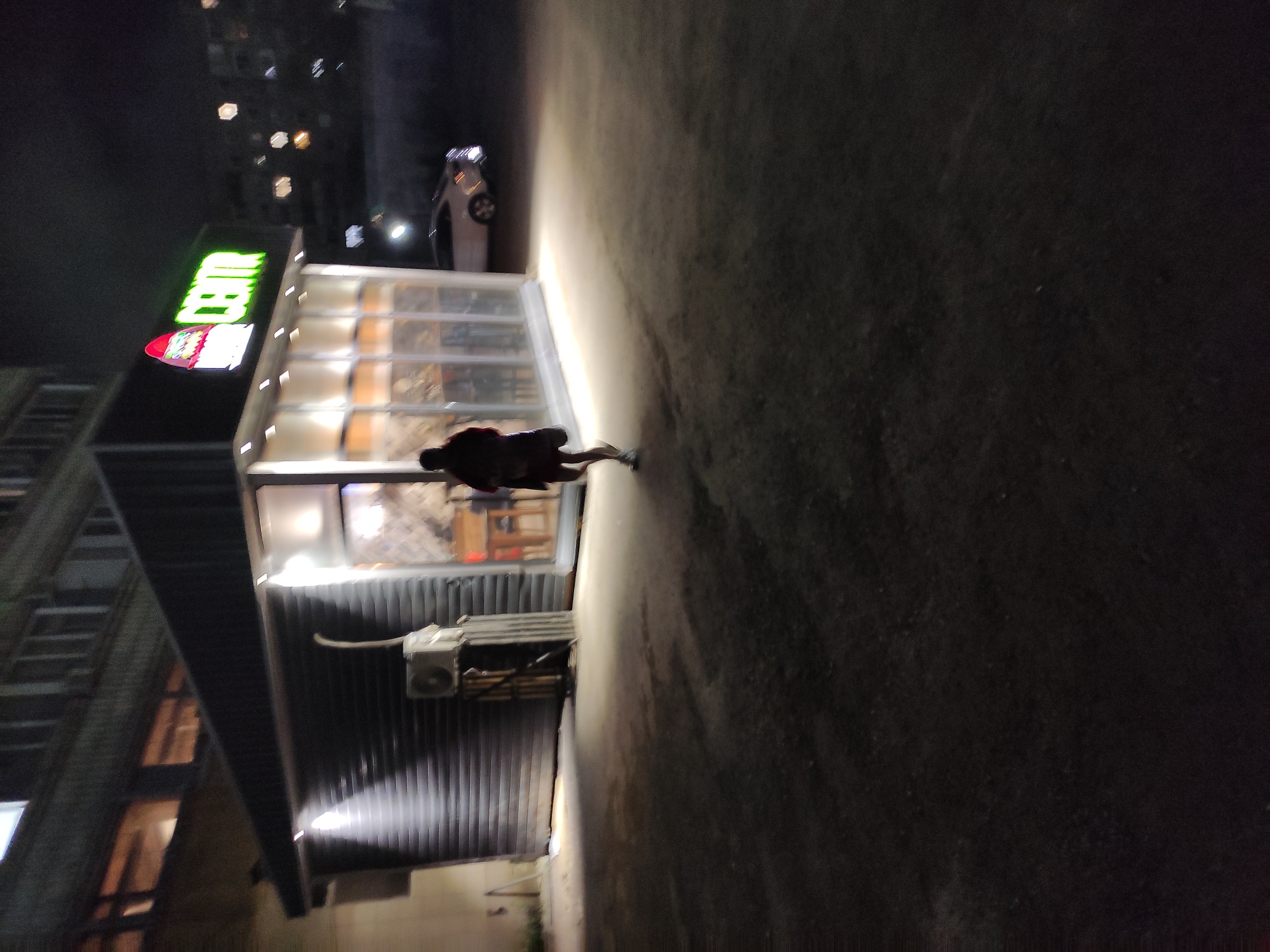 A night scene in Pavlodar, Kazakhstan, shows a building with a brightly lit store with the word "CENTR" visible on the signage. A person is walking on the sidewalk in front of the store. A parked car is visible behind the building. The surroundings suggest an urban setting with buildings and streetlights.