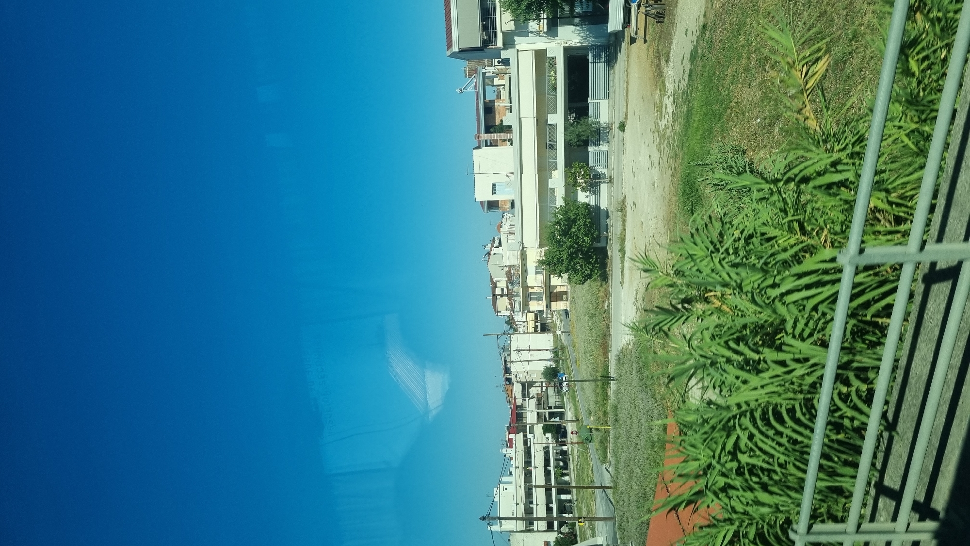 The image captures a view from a moving vehicle, likely a bus or car, looking towards a suburban area under a clear blue sky. In the foreground, lush green reeds and a metal railing suggest the perspective is from a roadway or bridge. Beyond this, a sparsely vegetated area gives way to a street lined with multi-story buildings. These buildings appear to be residential or commercial, with a mix of white and cream facades, balconies, and some with awnings. Power lines and poles are visible, indicating typical urban infrastructure. Further in the background, more buildings are visible, some with red tiled roofs. A light blue vehicle is parked on the street, and a green trash bin is also present. The overall scene depicts a sunny day in a residential district of Larissa, Greece, with the characteristic architecture and infrastructure of the region.