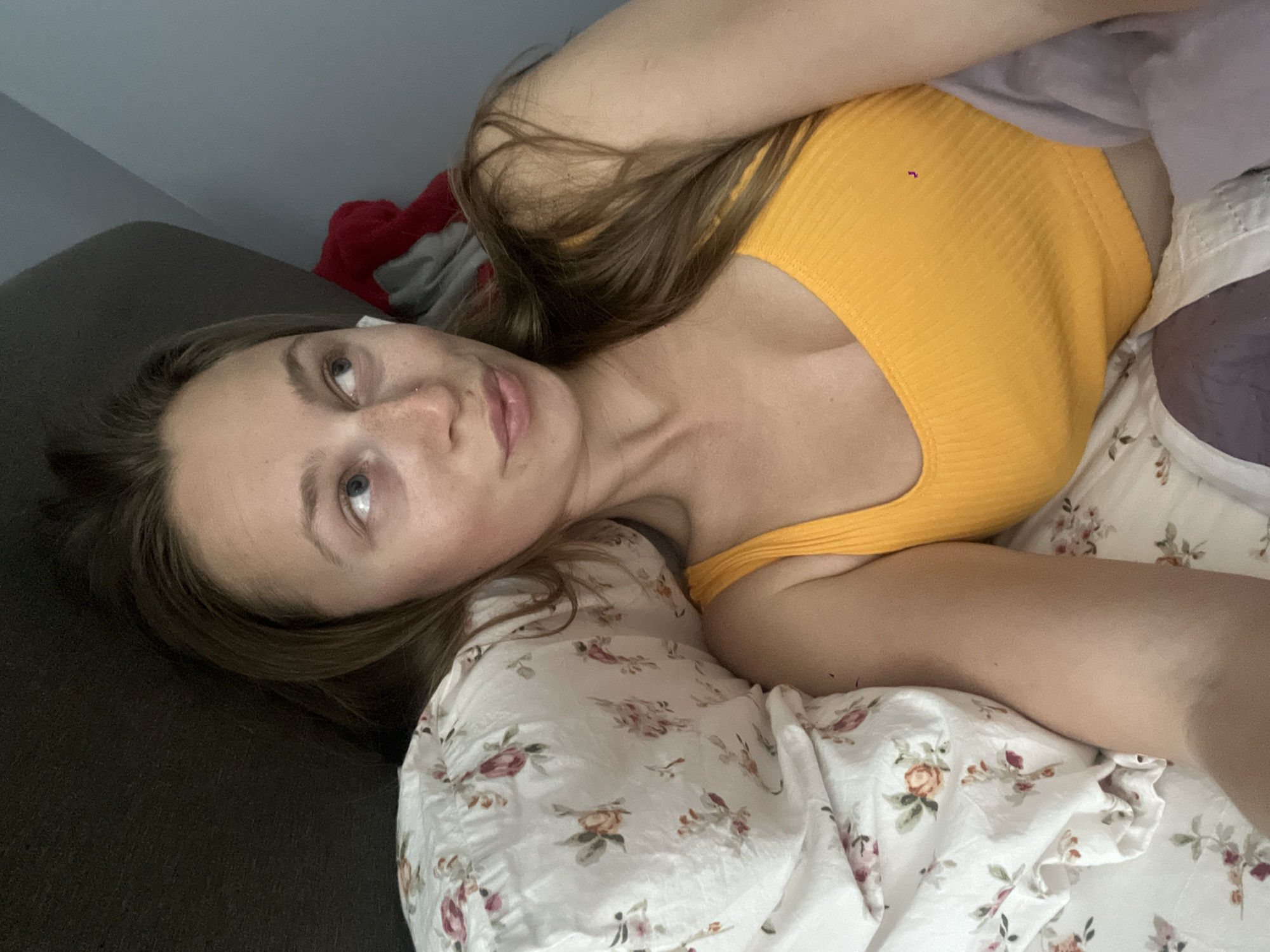 The image depicts a single female subject in an indoor setting located in the city of Bečići, Montenegro.
**Subject:** A Caucasian female with long, light brown hair, blue eyes, and light freckles across her nose and cheeks. A small stud piercing is present on her left nostril. She is wearing a yellow, ribbed, scoop-neck tank top.
**Action and Position:** The female is lying on her left side, head resting on a pillow. Her left arm is partially visible, extending towards the camera, suggesting a self-portrait. Her gaze is directed upwards and to the right within the frame, with her lips slightly parted and pursed in a contemplative expression.
**Environment and Objects:**
* **Background:** A dark brown or charcoal gray upholstered surface is visible on the left side of the frame, likely a headboard or couch. A light blue-gray wall occupies the upper right background, featuring a visible corner or ceiling line.
* **Bedding:** Her head rests on a white pillow adorned with a floral pattern of roses and leaves in shades of red, pink, and green. A light purple or lavender blanket or sheet covers the lower portion of her torso.
* **Miscellaneous:** In the upper right corner, against the wall, partially visible crumpled red and gray fabrics are present.
* **Lighting:** The scene is illuminated by soft, diffuse lighting, consistent with an indoor environment.