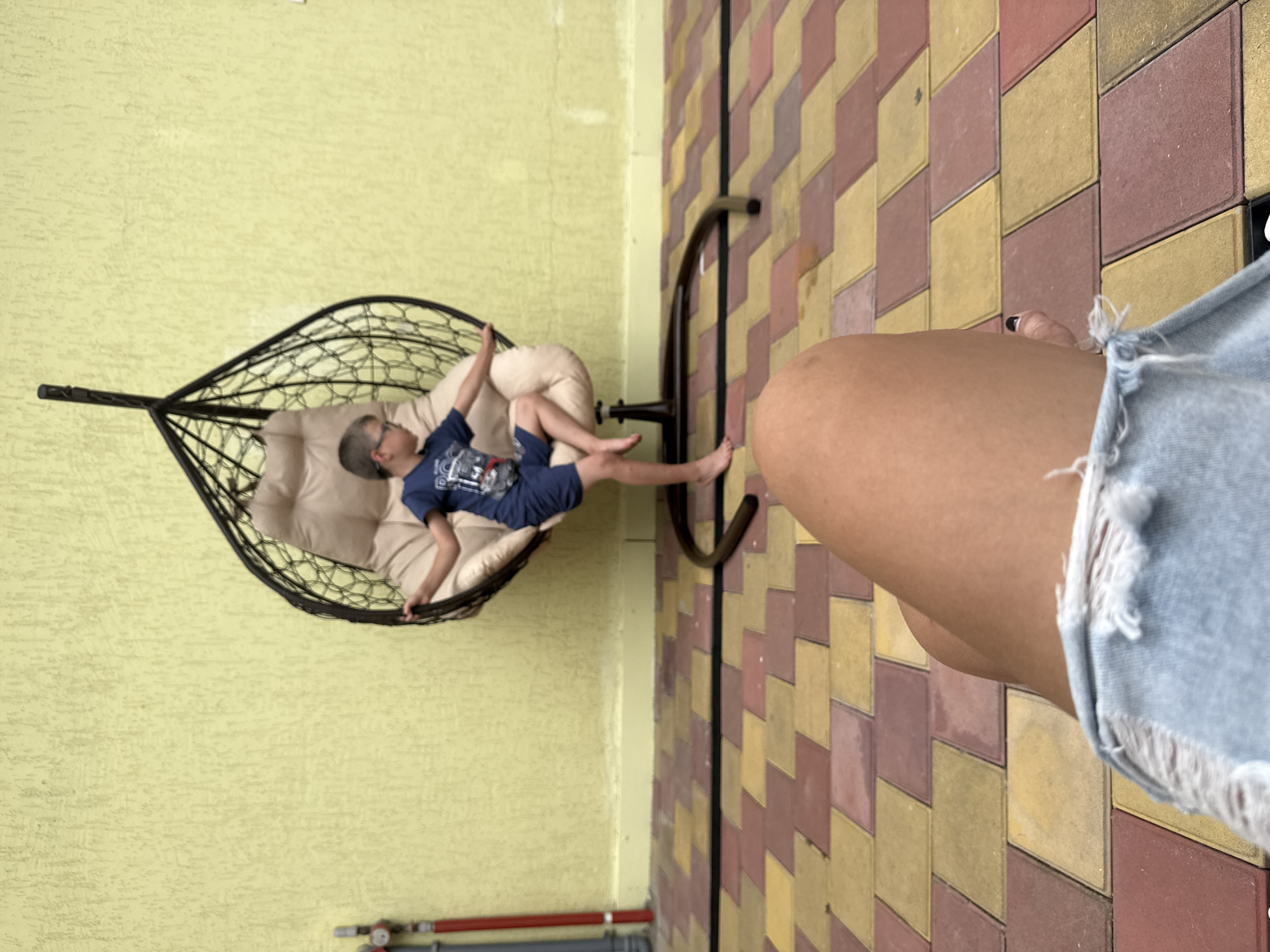 In a courtyard with a yellow textured wall and a multi-colored paver tile floor, a young boy wearing glasses, a blue t-shirt, and blue shorts sits in a hanging egg-shaped chair. He is looking to his right and has his right leg crossed over his left. The chair is dark brown with a beige cushion. In the foreground, a person's bare leg and torn denim shorts are visible, with a manicured fingernail on the right hand. A red and gray pipe assembly is mounted on the left side of the wall. The scene appears to be outdoors, possibly in the city of Sirius, Russia.