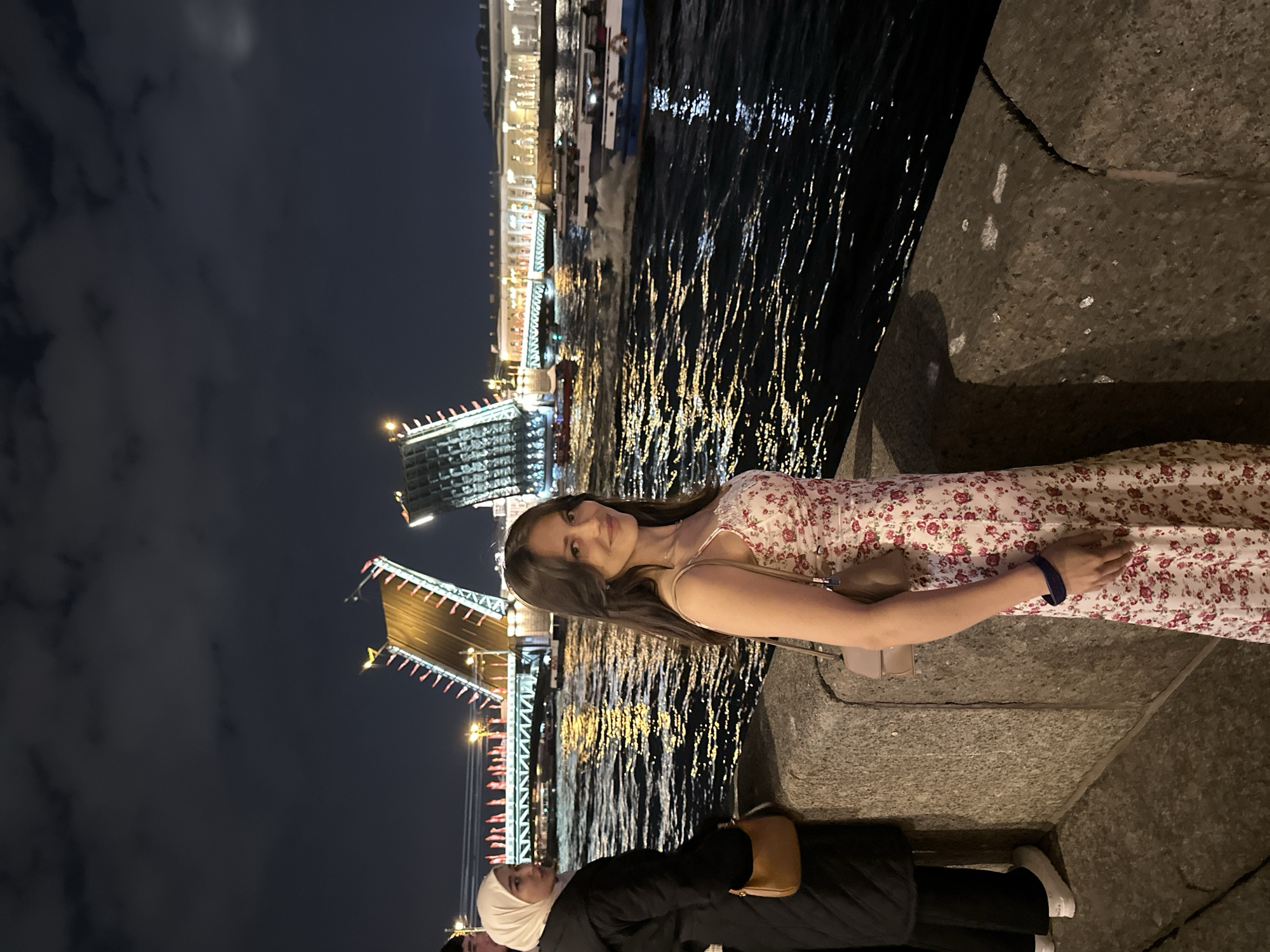 At night in Saint Petersburg, Russia, an open bridge stands over a body of water with reflections. A person in a floral dress and a shoulder bag leans against a concrete ledge. Below, another person wearing a black outfit and white headscarf sits.