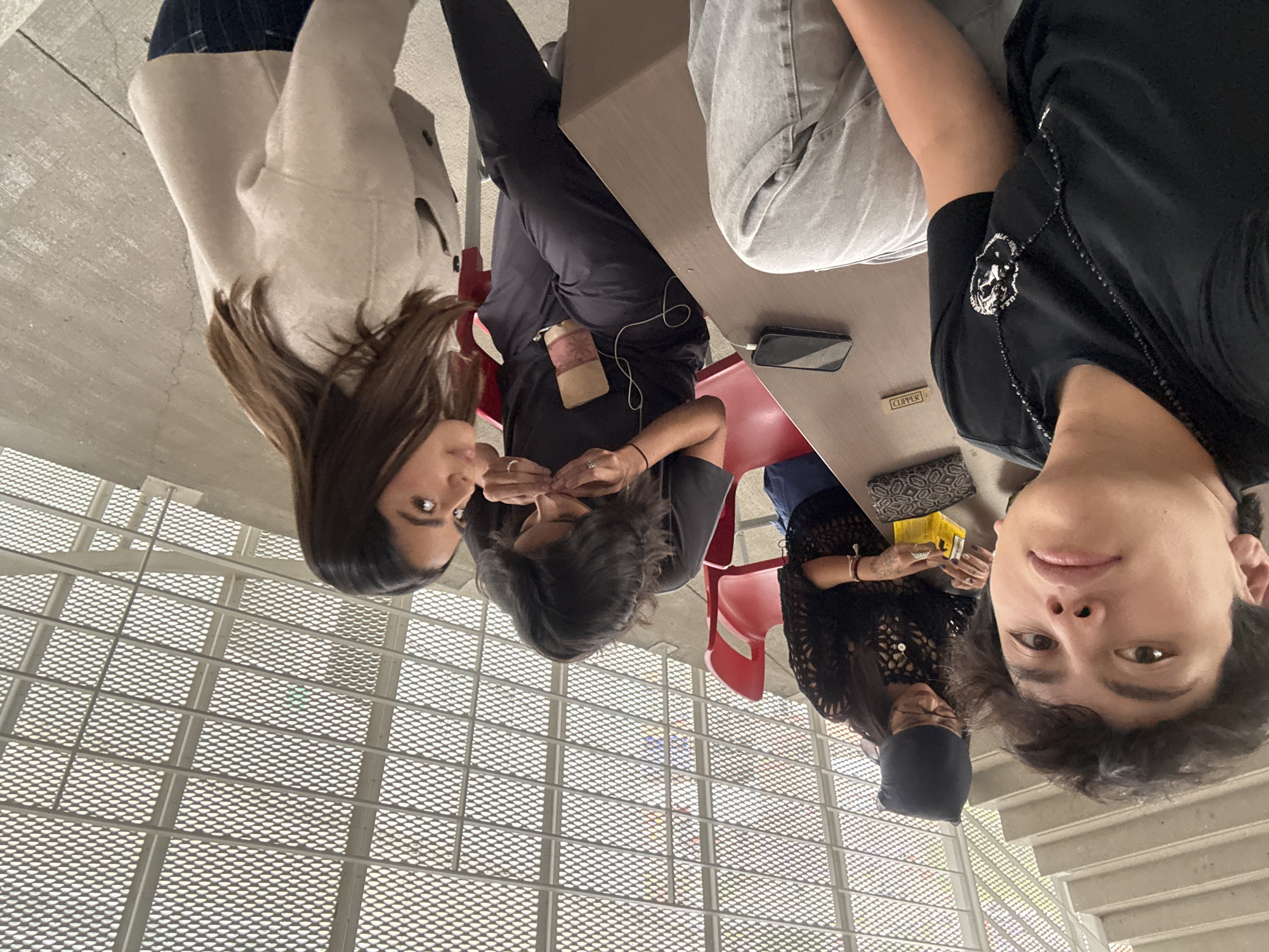 Four young adults are pictured in a casual indoor setting, likely a modern building such as a university or public area, given the distinctive grid-patterned ceiling and visible stairs. The perspective is from a slightly low angle, looking up at the individuals who appear to be seated or leaning around tables.
In the foreground on the right, a person with short, dark, curly hair looks directly at the camera with a neutral to slightly smiling expression. They are wearing a black short-sleeved top adorned with a silver and black pendant on a beaded necklace. Adjacent to them, on the left, another individual is seen wearing a black, open-knit or crocheted-style top and a black head covering. They are looking down at a table where a yellow box, a small patterned pouch or wallet, and a "CLIPPER" branded lighter are visible.
Further to the left, a third person with dark hair is also looking downwards, seemingly focused on an object in their hands, possibly a phone with white wired earphones connected, or adjusting a light brown pouch attached to their black attire. On the far left, a person with long brown hair, dressed in a light-colored long-sleeve top and dark pants, is looking towards the center of the group with a gentle expression.
The group appears to be engaged in a relaxed social gathering or rest period. Light-brown tables and hints of red chairs are present. A black mobile phone is also visible on one of the tables. The background features a striking light-grey, grid-patterned ceiling or floor structure of an upper level, suggesting contemporary architecture. The scene is illuminated by natural daylight.