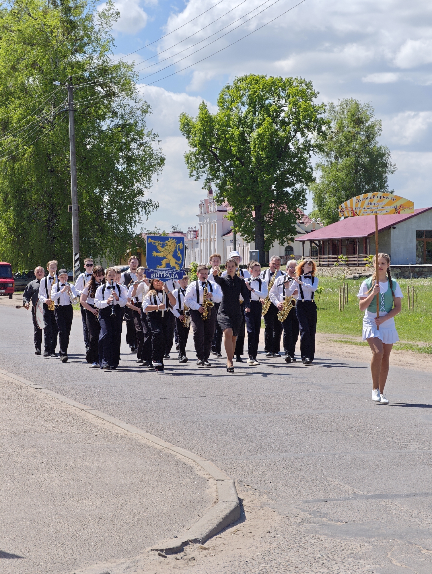 The image captures a marching band parading down a street in Парэчча, Belarus. The band members, primarily young individuals, are dressed in formal attire, consisting of white shirts, black pants or skirts, and suspenders. They are playing various instruments such as clarinets, saxophones, and percussion. At the head of the band marches a woman in a black dress and heels, leading the group. A young girl in a white skirt, white sneakers, and a green vest walks on the far-right of the frame, carrying a sign. A large banner with a heraldic lion and the word "ИНТРАДА" is carried by band members.
The parade takes place on a paved road with a curb on the left side. Trees and buildings form the backdrop. A utility pole with power lines is visible on the left. A building with a red roof and a sign reading "ДУХОВОЙ ОРКЕСТР" is visible in the background. The sky above is partly cloudy. A red car is parked on the left side of the frame.
