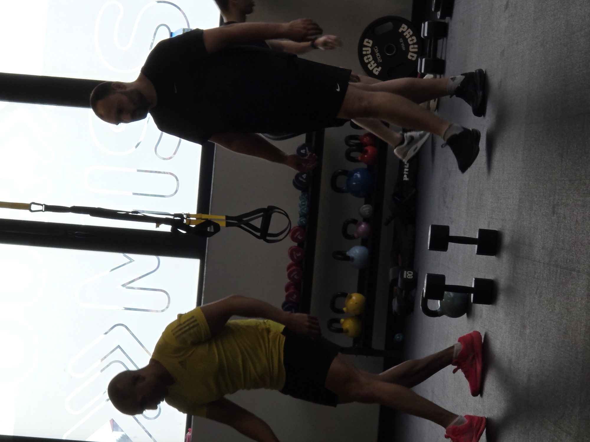 The image depicts two men engaged in exercise within a gym or fitness studio, illuminated by strong backlighting from a large window. The scene is set indoors, likely during the daytime.
In the foreground, a man wearing a yellow short-sleeved shirt, black shorts, and distinctive pink/red athletic shoes is positioned as if performing a plank or similar bodyweight exercise, with his body held straight and his gaze directed downwards.
Behind him, another man dressed in a black short-sleeved shirt, black shorts with a small white Nike swoosh logo, and black/white athletic shoes stands in an athletic posture, also looking down.
The gym environment is equipped with various fitness apparatus. A rack holding an assortment of kettlebells in red, blue, and yellow hues is visible. Nearby, dumbbells are placed on the dark gray floor, along with a 20-kilogram weight plate, which clearly displays the brand name "PROUD" and "20KG". A TRX suspension trainer is also hanging in the upper left portion of the image.
The prominent window behind the men features large, stylized white outlines of letters, though no complete word or phrase is fully discernible. The backlighting creates a strong silhouette effect, obscuring detailed facial features or expressions of the individuals. The overall ambiance is that of a functional training space.
