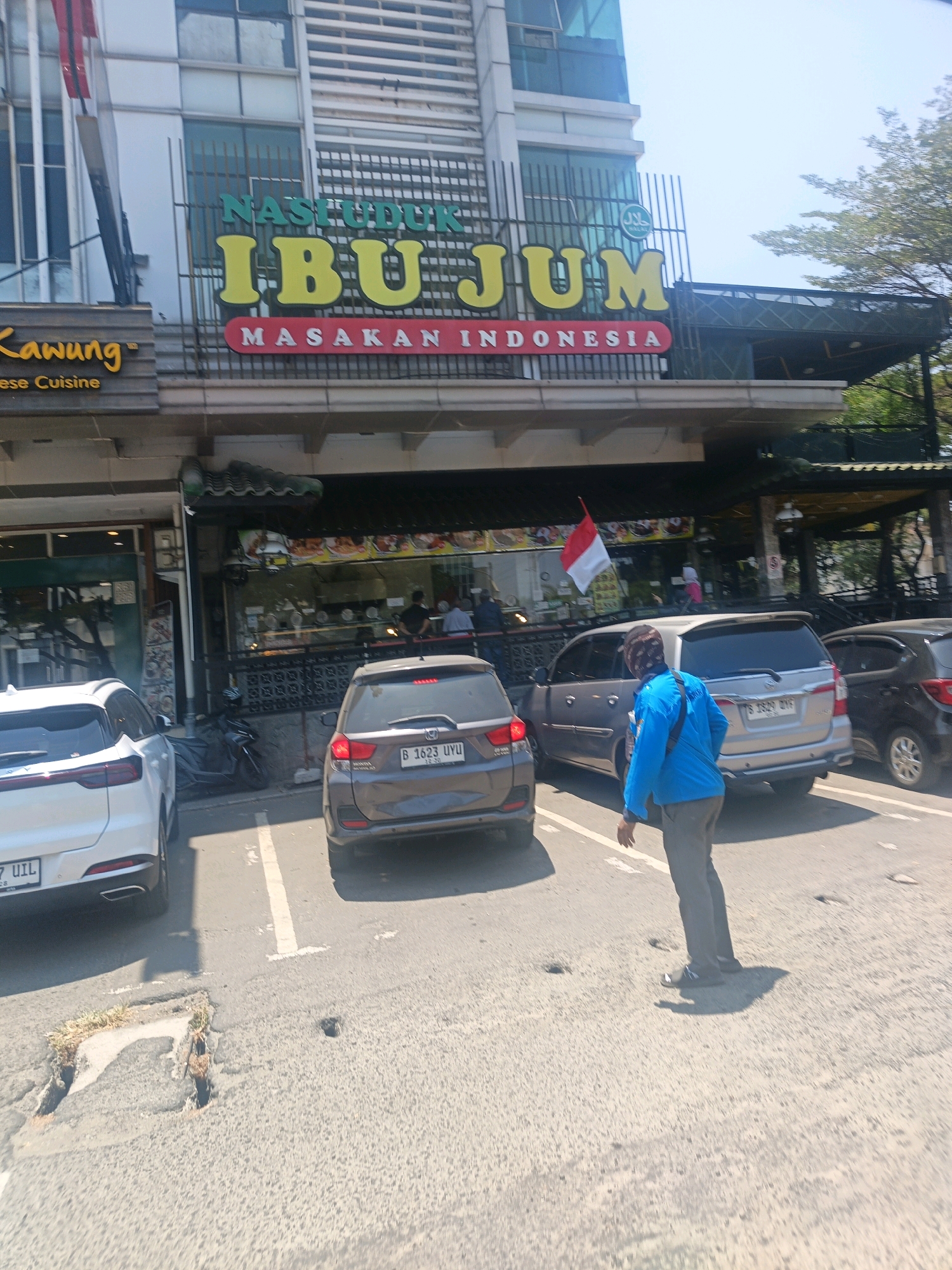 A bright, daytime, eye-level, medium shot captures the exterior of a restaurant in North Jakarta, Indonesia. The restaurant, identified by the prominent signage "NASI DUK IBU JUM" in large yellow letters and "MASAKAN INDONESIA" in red below, features a dark green awning extending over a food display area visible through glass windows. To the left of the restaurant, a separate establishment has a wooden facade with the name "Kawung" and "nese Cuisine" visible. In front of the restaurants, several cars are parked in designated spots on a paved surface. A dark grey Honda Mobilio is parked center-right, with its license plate B 1623 UYU. To its left is a silver Toyota Innova. On the far left, the rear of a white SUV is visible, with its license plate partially obscured. A person wearing a blue jacket and grey pants is walking towards the right of the frame, casting a shadow on the pavement. A small red and white flag is attached to a pole above the restaurant's awning. The sky is clear and blue, with the top of a tree visible on the right side of the image. The overall scene depicts a typical commercial street environment.