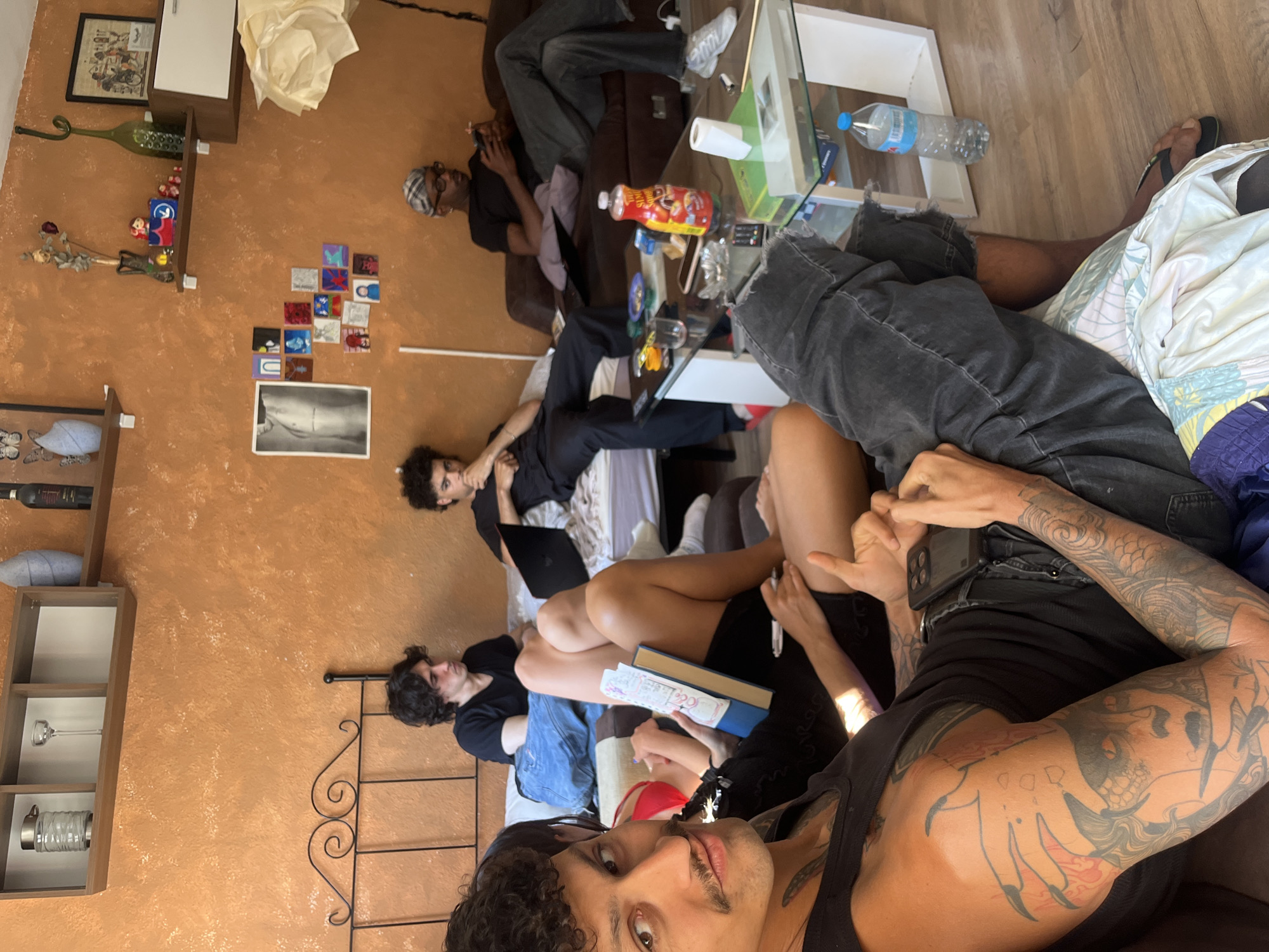 The image depicts a room in Barcelona, Spain, with a group of people lounging. One person in the foreground is taking a selfie, showing tattoos on their arm and wearing a black tank top. Several others are lying on a sofa, engaged in various activities like reading, using laptops, or relaxing. A glass coffee table is visible, holding snacks, drinks, and personal belongings. The room's decor includes artwork on the wall, decorative objects on a shelf, and various pieces of furniture. The atmosphere seems relaxed and casual.