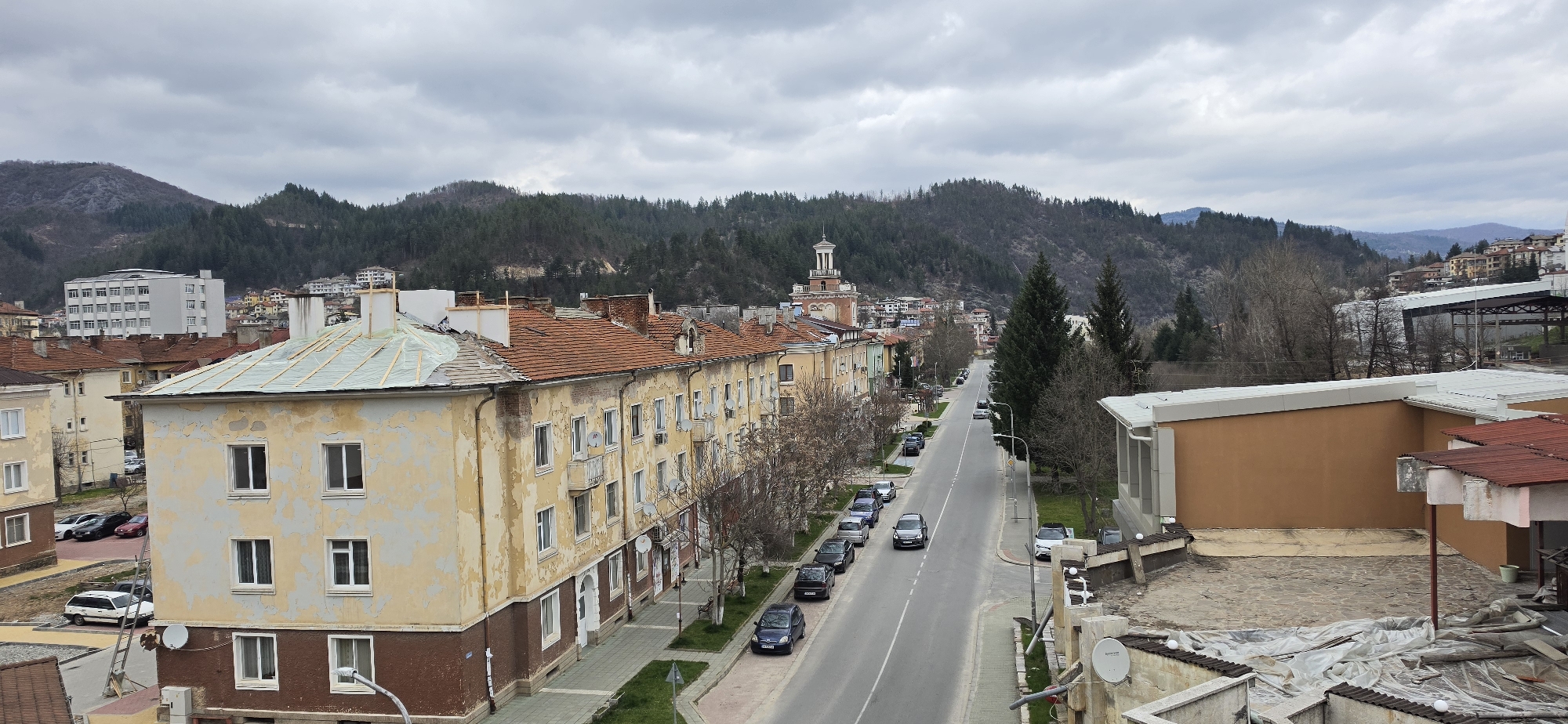 This is an elevated outdoor shot of a street in Rudozem, Bulgaria, taken on an overcast day. The street, with a central double yellow line, runs away from the viewer into the center of the frame. There are several parked cars alongside the street on both sides. The buildings flanking the street are older, multi-story residential structures with peeling, weathered exteriors in shades of beige and brown. Many have balconies with satellite dishes. The roofs are primarily red tile. The building closest to the viewer on the left has a green tarp-covered roof. In the background, a line of pine-covered mountains dominates the skyline. A tower structure with a white cupola and orange brick exterior stands prominently in the middle distance. A newer building is visible on the right. There are sidewalks and trees lining the street. No people are visible in the shot.
