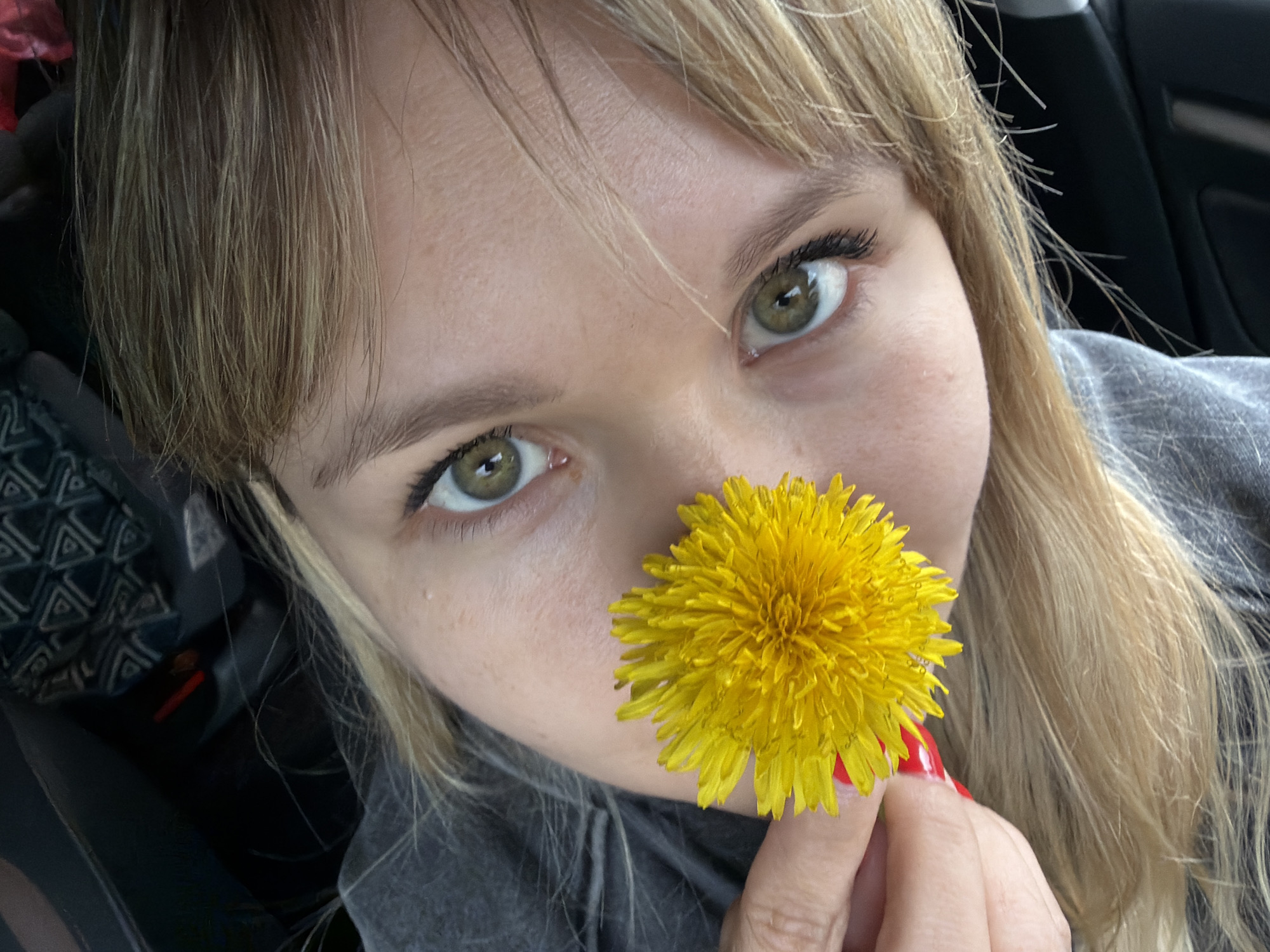 A close-up selfie of a young woman with light blonde hair and green eyes. She is holding a bright yellow dandelion in front of her nose. The background is blurry and dark, suggesting she is inside a car. The location is Chaikovsky, Russia.
