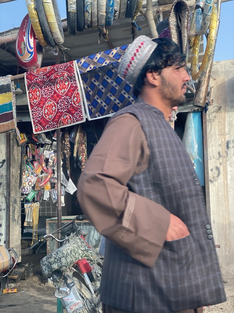 In Mir Ahmad Kalay, Afghanistan, a man with a beard and wearing a traditional pakol hat, a brown tunic, and a dark plaid vest walks past a shop. The shop's roof is adorned with tires and colorful hanging textiles. Various trinkets and motorcycle parts are displayed within the shop's confines. A camouflaged motorcycle is parked in front of the shop.