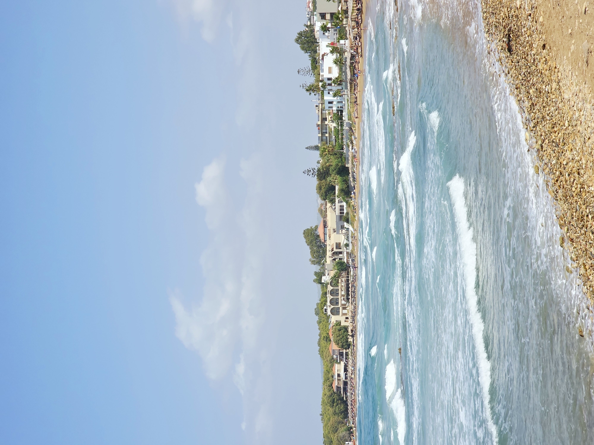 This is a vertical, eye-level shot of a beach scene in Daratsos, Greece. The foreground is dominated by a pebble beach, transitioning into a sandy area where numerous beach chairs and umbrellas are set up. Beyond the beach, the turquoise ocean is active with white-capped waves. In the mid-ground, a line of buildings, appearing to be hotels and restaurants, stretches along the coastline, backed by a dense green forest. The sky above is a clear light blue with a few scattered white clouds. The scene is bathed in daylight, suggesting a warm, sunny day. There are no people clearly visible as distinct individuals, but their presence is implied by the organized beach furniture and the distant structures suggesting an inhabited area.