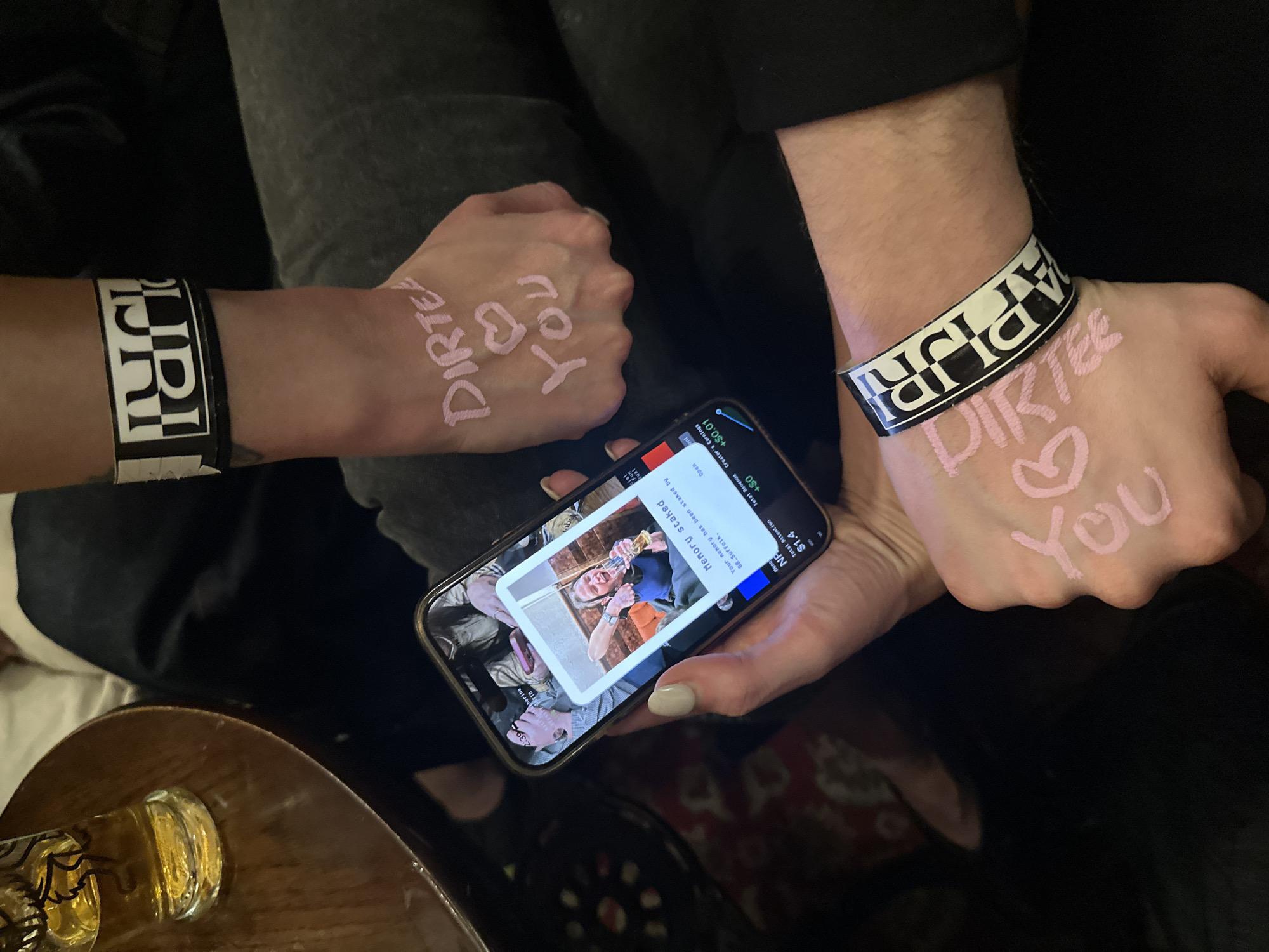 Two people are seated indoors, displaying their hands. Each wears a black and white wristband labeled "DIRTEE," and pink writing on their hands spells out "DIRTEE <3 YOU." One person holds a smartphone displaying a picture of another person toasting a drink, with a notification indicating "Memory staked," earning $0.01 for the creator and generating $1.4 in total attention. A glass containing a liquid is visible on a nearby table. The OCR suggests the picture’s content creator, referred to as “68.Suffolk”, is located in the city of London.