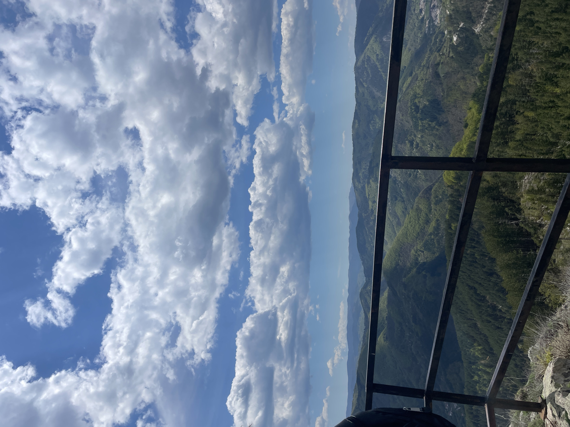 The image shows a mountainous landscape from an elevated viewpoint in Gradat, Bulgaria. A metal railing is in the foreground. Behind the railing is a view of green, forested mountains receding into the distance. The sky is blue with puffy white clouds.