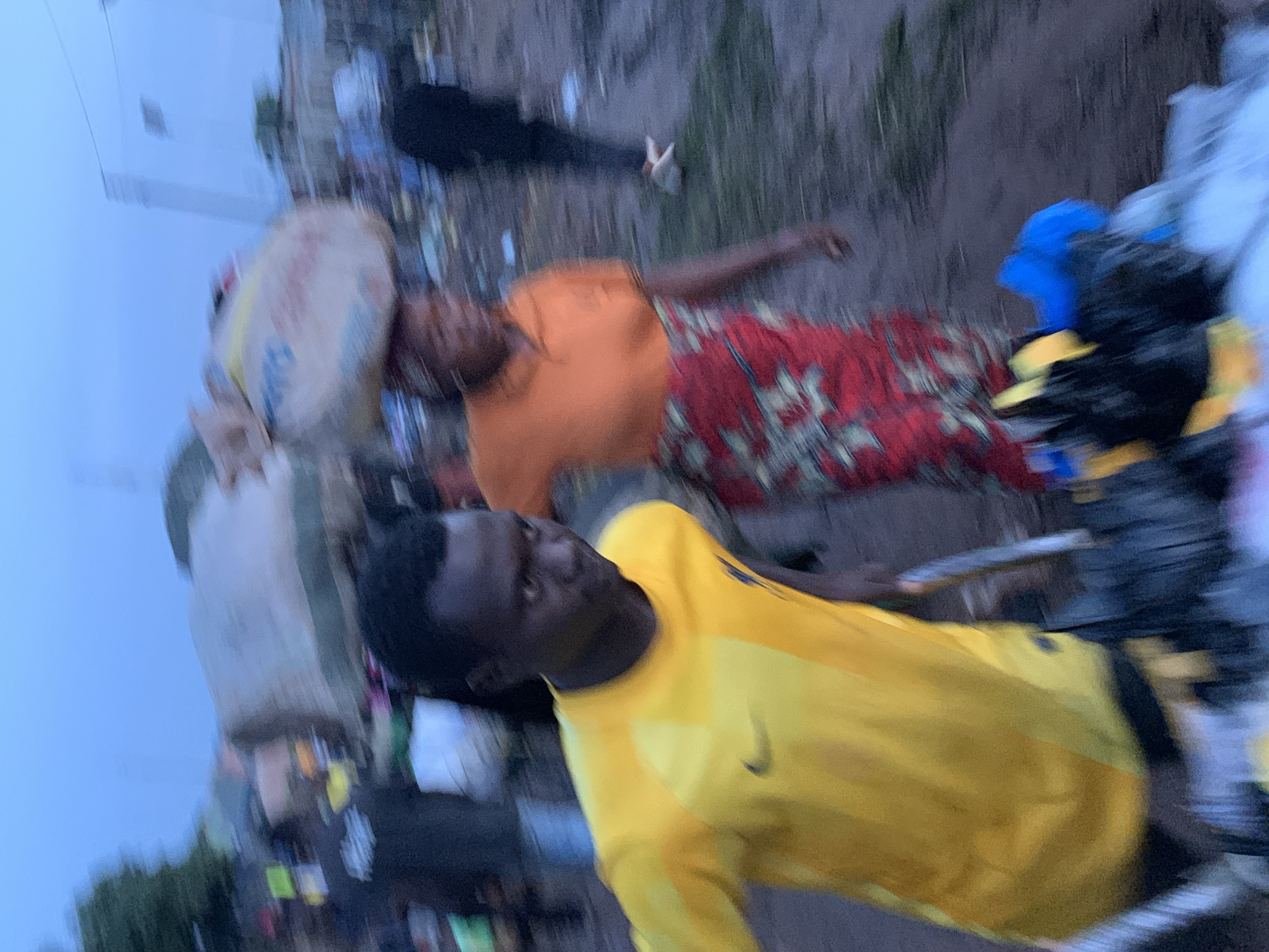 In the city of Gura Top, Nigeria, a blurred scene captures a marketplace atmosphere. A young man in a yellow t-shirt is in the foreground, looking to his right. Behind him, a woman with a large bag on her head, wearing an orange shirt and a red and green patterned skirt, walks away from the camera. In the background, other figures and structures are indistinct due to motion blur. The environment appears to be an outdoor, dusty, and possibly crowded area.