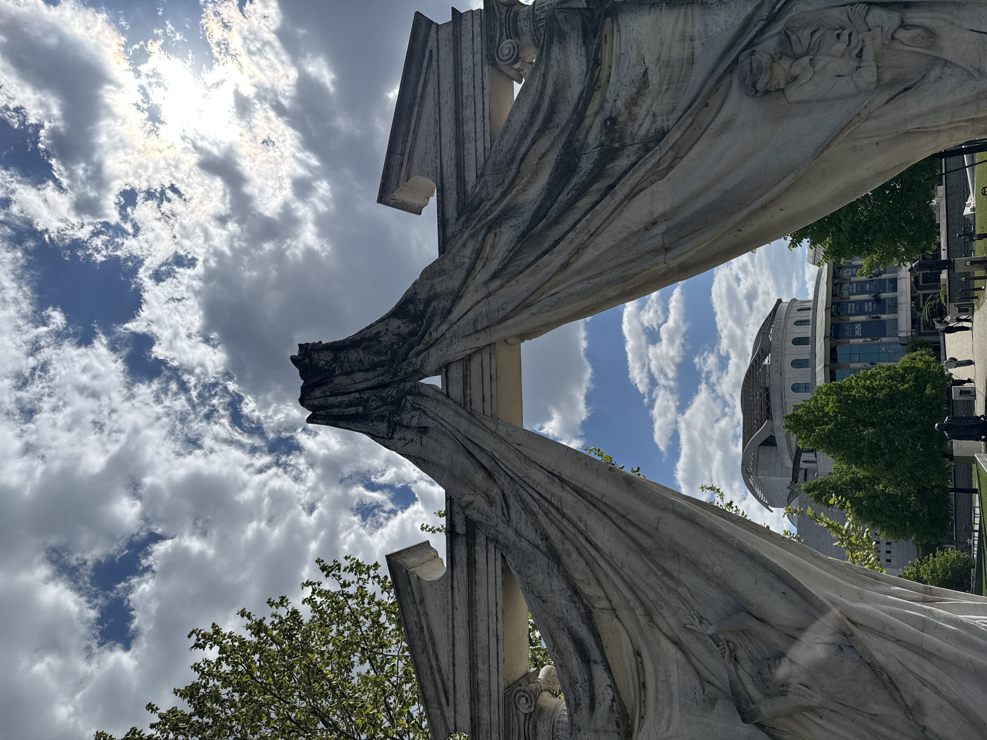 The image presents a low-angle view of the "Nemzeti Színház" or the National Theatre, framed by the drapery of a monumental stone structure, likely part of a memorial or monument. The structure is composed of stone pillars and detailed carved drapery. The sky is filled with a mix of partly cloudy and blue patches. The theatre, in the background, appears to be a modern architectural design with large glass windows. The foreground contains hints of green foliage, suggesting a park or garden setting. People are walking in front of the theatre. The location is Budapest, Hungary.
