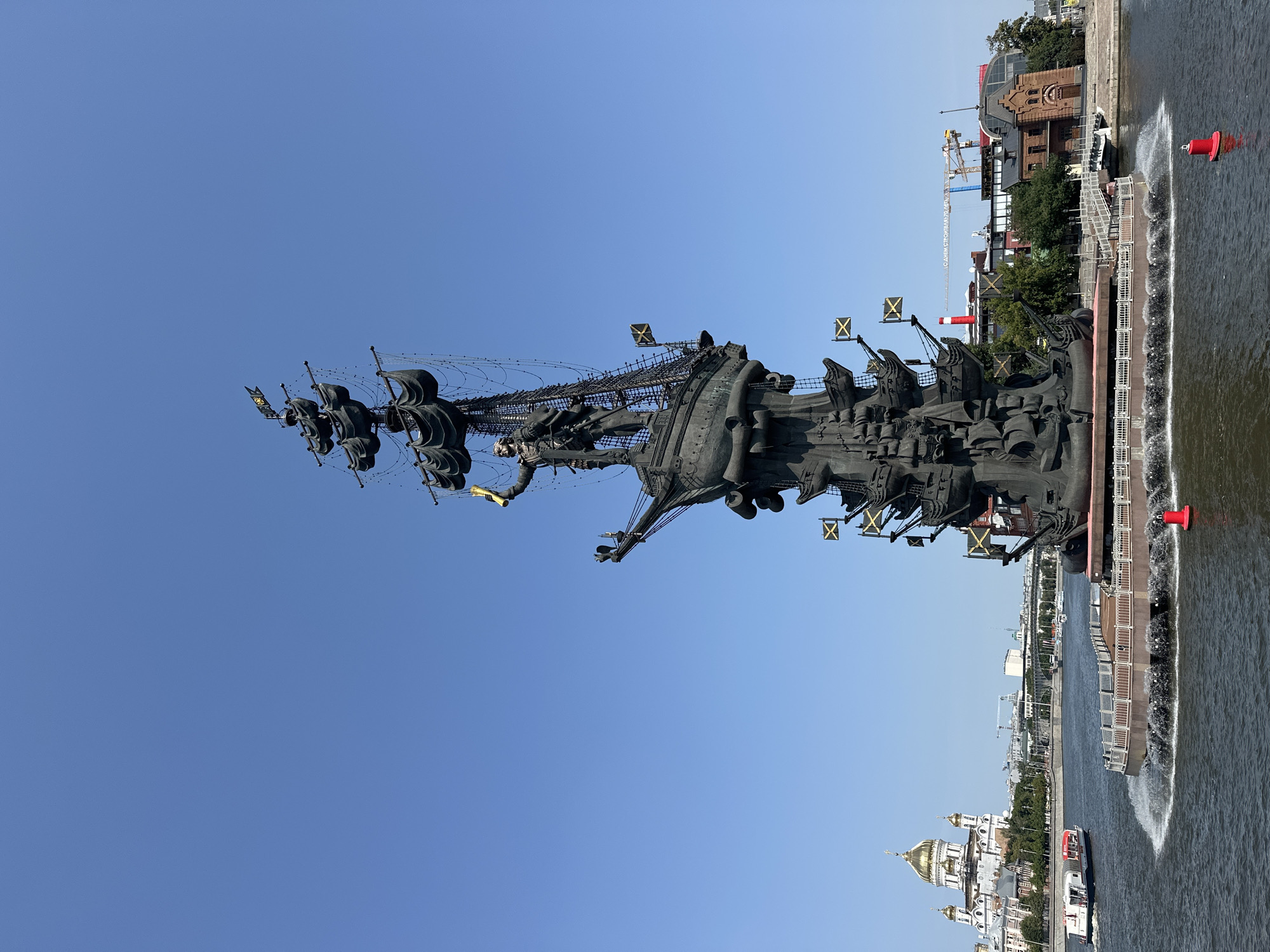 The image shows the Monument to Peter the Great in Moscow, Russia, on a clear, sunny day. The massive bronze monument, depicting Peter the Great standing on a ship with multiple masts and sails, dominates the foreground. The monument is situated on a circular platform in the Moskva River, with water spraying around the base. In the background, across the river, is the Cathedral of Christ the Saviour with its distinctive golden domes, and further in the distance, a cityscape with buildings and cranes. Several boats are visible on the river, including a white tourist boat with red accents moving to the left. The sky is a vibrant, cloudless blue. The monument is adorned with several flags, some bearing a yellow cross on a black background. The overall scene is a majestic and iconic view of a significant landmark in Moscow.